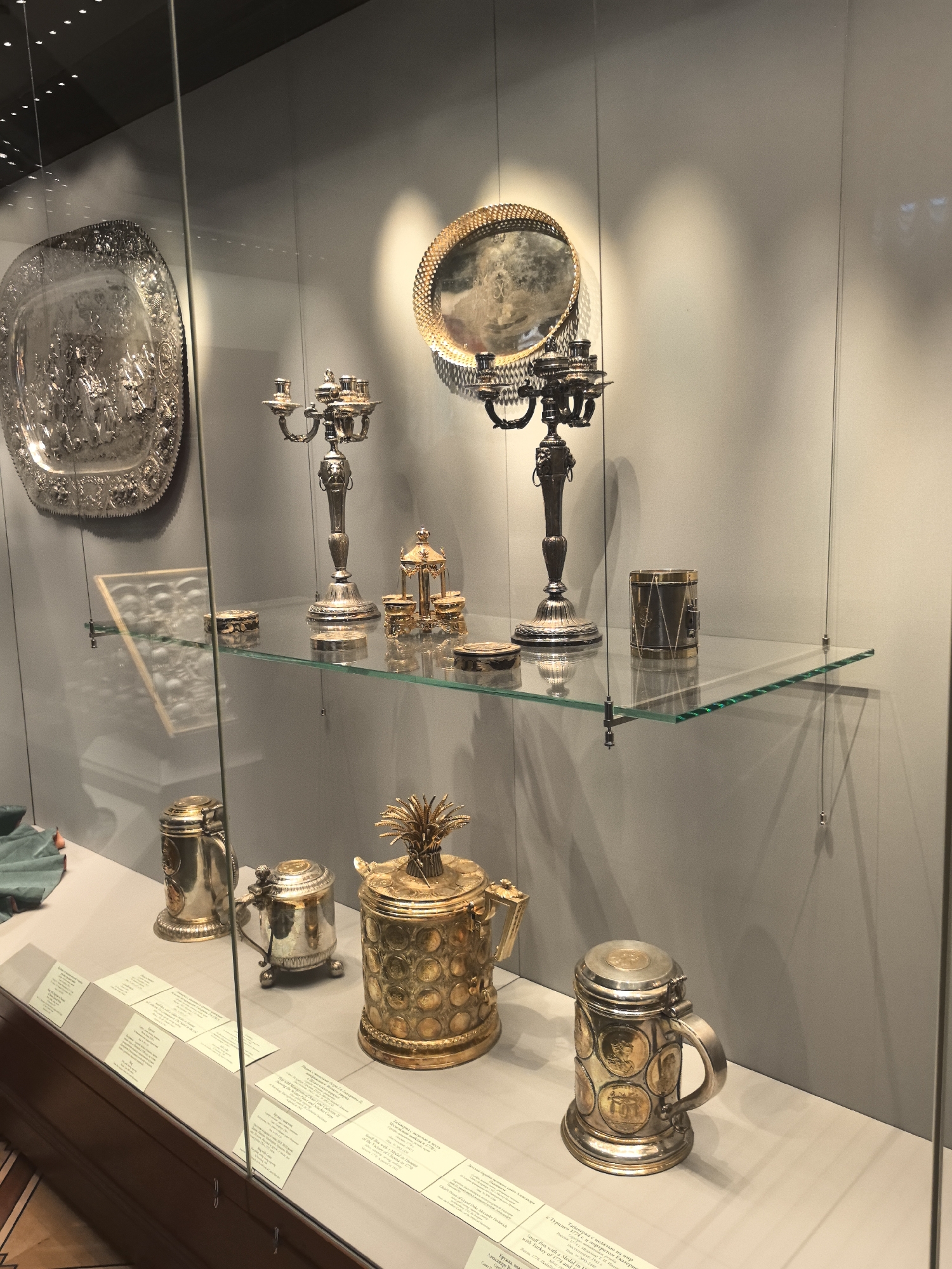 This media file shows a display case at the State Hermitage Museum in Saint Petersburg, Russia. The display features an assortment of antique silverware and goldware, presented on a glass shelf and a flat surface behind it. Among the items are ornate candelabras, a large decorative silver platter with intricate relief work, and several lidded vessels, including a striking gold pitcher topped with a pineapple finial. Also visible are two silver tankards, one adorned with circular medallions. The lighting suggests an indoor museum setting, with spotlights highlighting the artifacts. Several descriptive cards, containing text in Russian and possibly English, are placed in front of the objects, providing information about their provenance and historical context. The overall scene conveys a sense of historical preservation and artistic appreciation. The text visible on one of the cards reads "Табакерка с медалью на мир с Турцией 1774 г. и портретом Екатери", which translates to "Snuff box with a medal on the peace with Turkey of 1774 and portrait of Catheri". This suggests that some of the displayed items are related to significant historical events and figures.