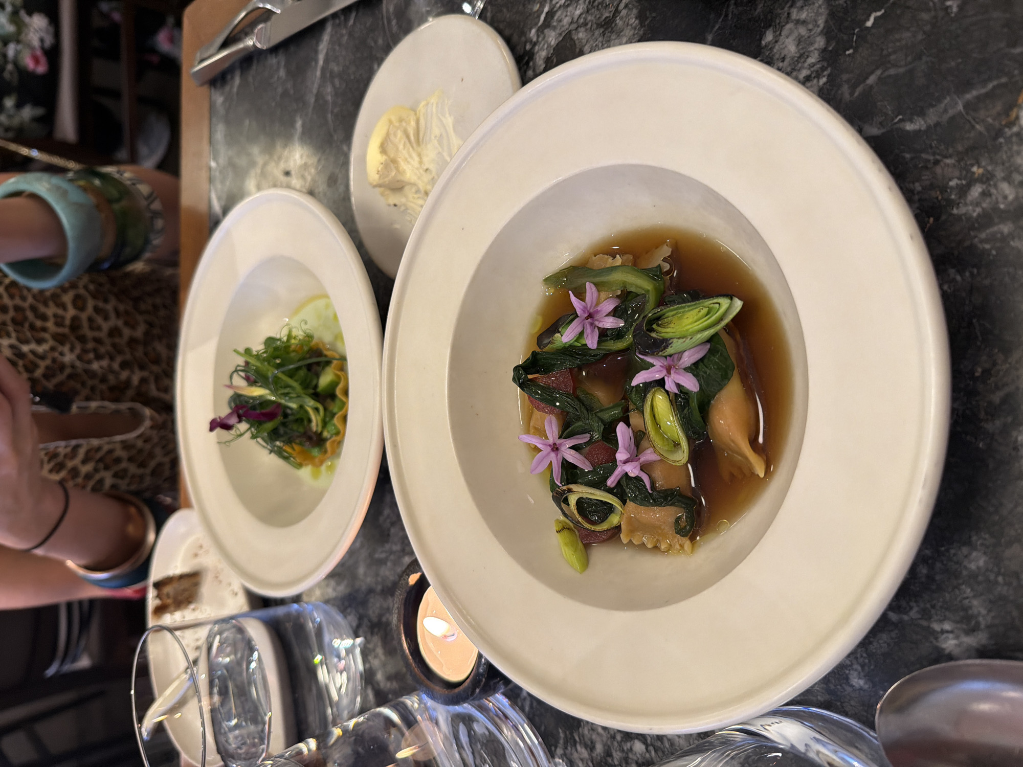 Two white ceramic bowls of food are on a dark marble table. In the foreground, a bowl contains ravioli in broth with green vegetables and purple flowers. To the left, a glass of water sits next to a small lit candle. Behind this, another bowl contains a small salad. A person's arm, wearing several bracelets, is visible in the background, and a fork is in the upper right corner of the frame. The setting appears to be a restaurant in London, United Kingdom.