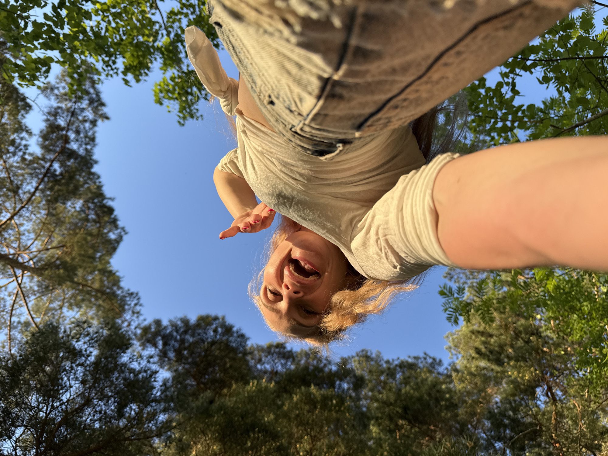 The image depicts a woman from a low-angle perspective, looking directly down at the camera. She is smiling with her mouth open and her hand is raised near her face. The sky is visible behind her, framed by tree foliage at the top and bottom of the image. The trees suggest an outdoor location, likely a park or wooded area. The blue sky indicates a sunny day. The woman is wearing a light-colored top and jeans. The image was purportedly taken in Прикамский, Russia.