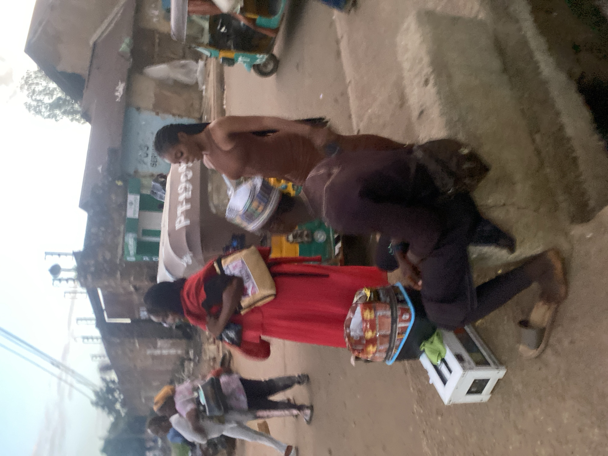 This street scene, located in Bukuru, Nigeria, captures a bustling environment under an overcast sky. In the foreground, a male individual wearing a patterned kufi cap, dark long-sleeved top, trousers, and sandals is seated on a light-colored concrete block on the right. To his left, a vendor's setup consists of a black plastic crate with a light blue rim, stacked with numerous cylindrical, foil-wrapped items. A small white wooden box with a green cloth rests on the ground beside the crate. Standing between this vendor setup and a light-colored three-wheeled vehicle (partially visible with "PT190" written on its side), is a female individual with braided hair, wearing a form-fitting brown sleeveless dress. She is oriented towards the left.
In the midground, a female individual in a long red dress is walking towards the left, carrying a light-colored handbag with a visible graphic design and a smaller dark bag. To her left, other pedestrians are in motion, including one person in light-colored pants and top carrying a dark bag, and another in a pink top. In the background, on the right, a turquoise and yellow three-wheeled auto-rickshaw is partially visible. The backdrop consists of several single-story buildings with corrugated metal roofs, indicating commercial or residential structures. One building has partial text "SER" and "203" or "703" on its facade, and another exhibits a green signboard. Overhead utility wires are visible against the sky in the upper left quadrant. The ground surface throughout the scene is unpaved and appears dusty.