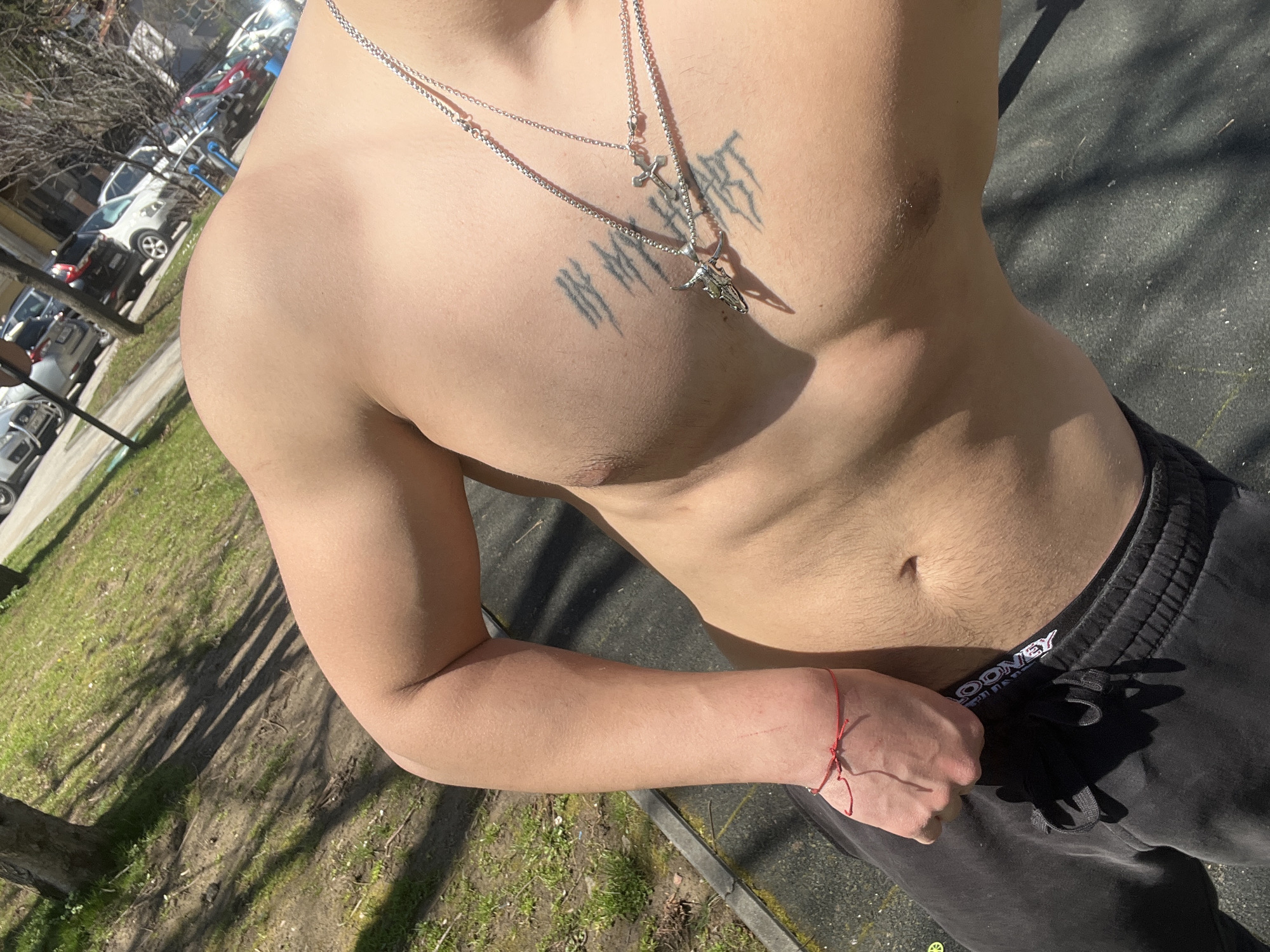 The image features a shirtless person with a fair complexion, wearing black sweatpants and two necklaces, one with a cross and the other with a bull skull pendant. A tattoo is visible on their upper chest. A red string bracelet is on their wrist. The background shows trees, grass, and parked cars, suggesting an outdoor setting, possibly in a park or near a residential area. The scene is brightly lit, likely due to sunlight. Based on the surroundings and the prompt, the image could be taken in Vratsa, Bulgaria, though the exact location cannot be determined without further visual cues.