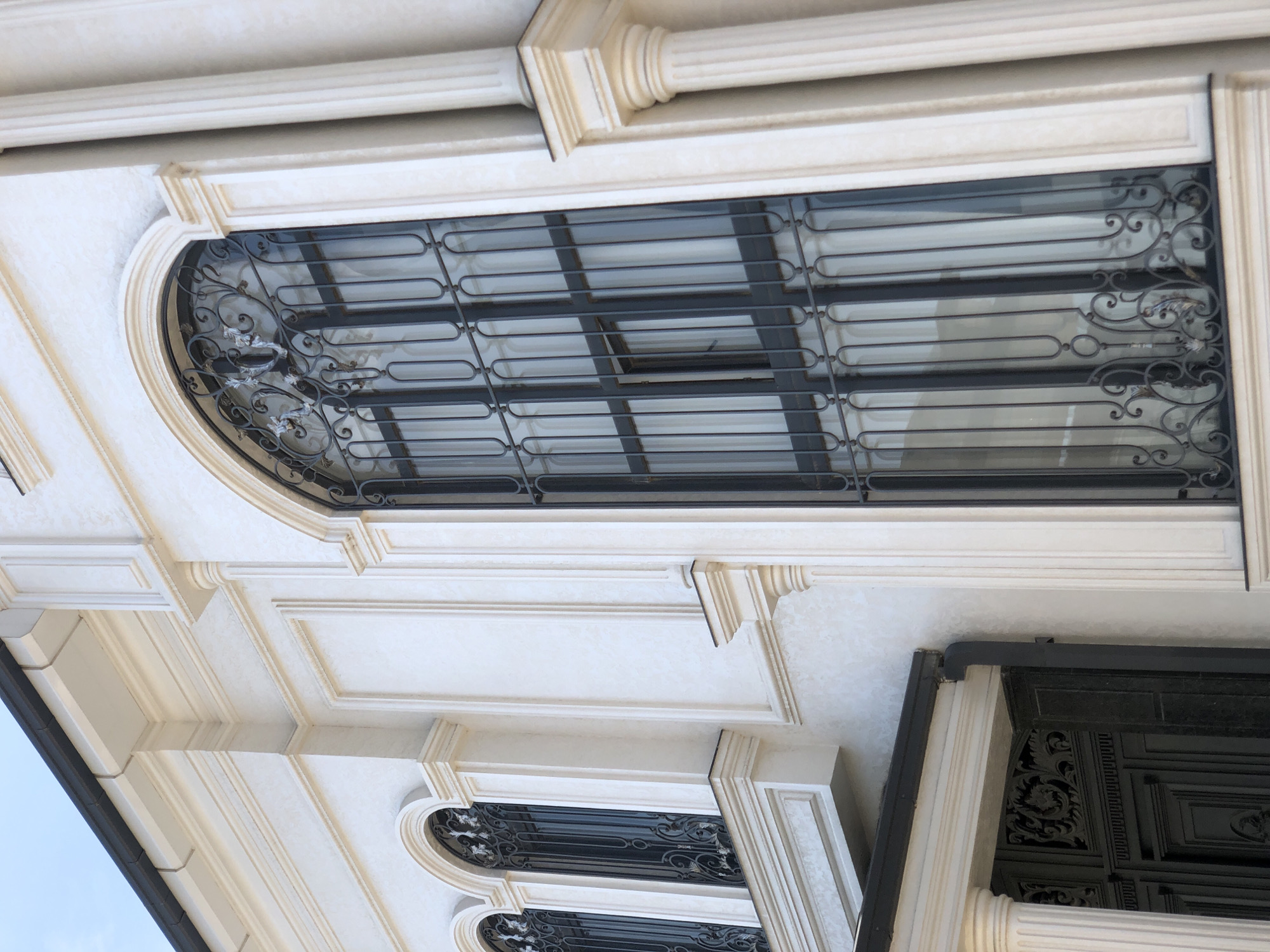 This image shows an exterior detail of an ornate building, likely a residence or public building, in Tashkent, Uzbekistan. The architecture features white stucco walls with decorative molding and cornices. Prominently displayed are large, arched windows with intricate black wrought-iron security bars and white sheer curtains visible behind them. Fluted pilasters and decorative capitals add to the classical aesthetic of the building's facade. A portion of a dark, ornate gate is visible at the bottom left, suggesting a grand entrance. The sky is a pale blue, indicating daylight. The overall impression is one of elegance and a well-maintained structure.