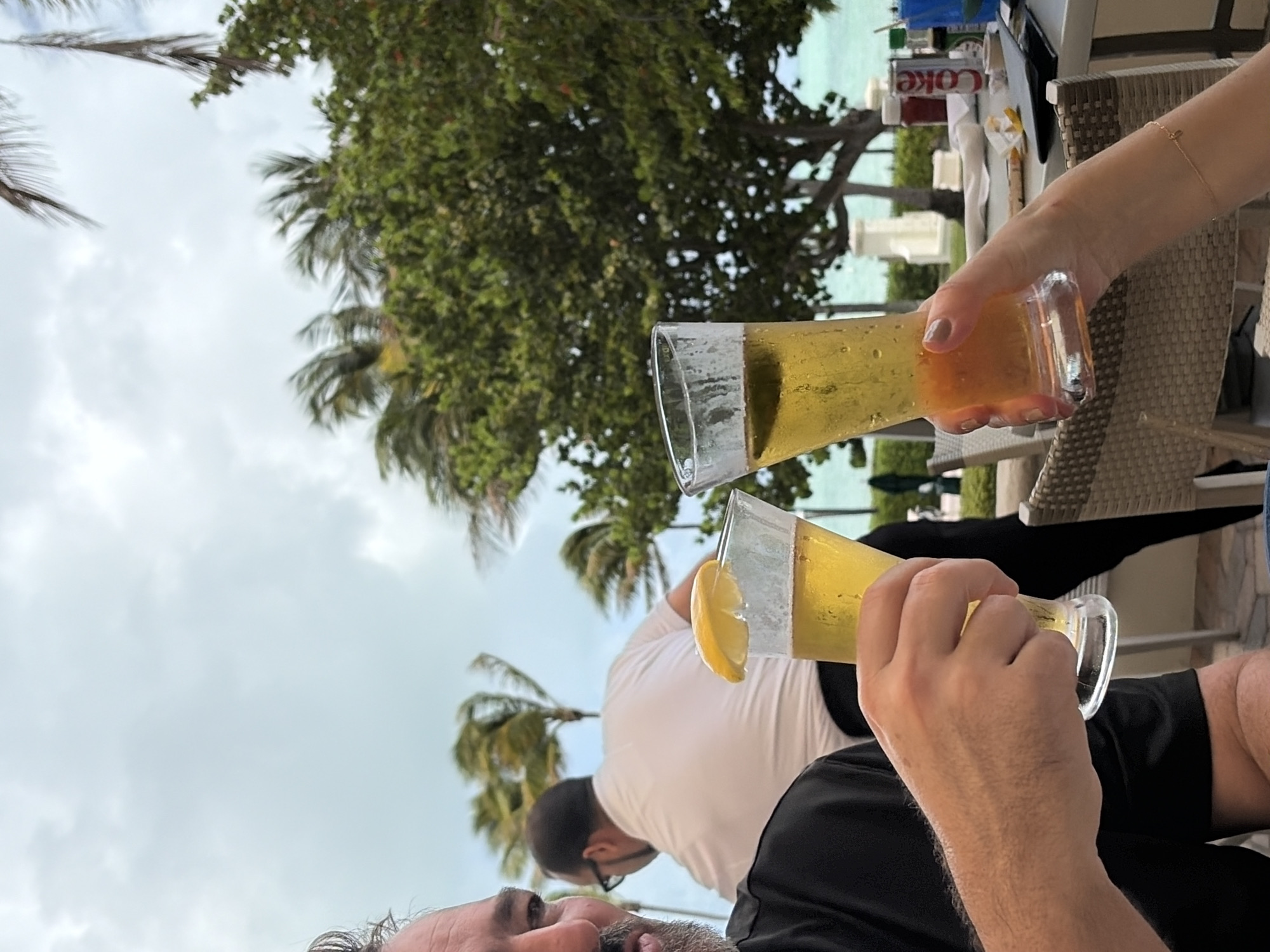 The image captures a convivial moment, likely a toast, in what appears to be an outdoor setting. Two individuals are raising glasses filled with yellow liquid, potentially beer or cocktails. One glass is adorned with a lemon slice. The backdrop includes lush vegetation featuring palm trees, suggesting a tropical locale. A glimpse of turquoise water hints at a coastal setting. Nearby, on a table, are beverage containers including a Coca-Cola can and other bottles. The scene seems to be captured from a low angle, likely from the perspective of one of the individuals involved in the toast.
Given the presence of palm trees and the turquoise water, as well as the fact that the image was taken in Key Biscayne, United States of America, the location is likely an outdoor restaurant or bar in the city.