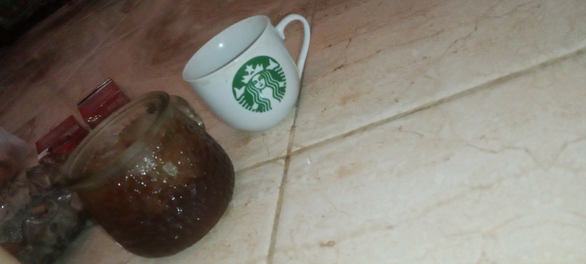 On a tiled floor, a white Starbucks mug with the iconic green siren logo rests next to a dark, round object that appears to be a coconut. Two small, rectangular red objects are also visible near the coconut. The lighting suggests an indoor environment. The scene's location is unspecified.