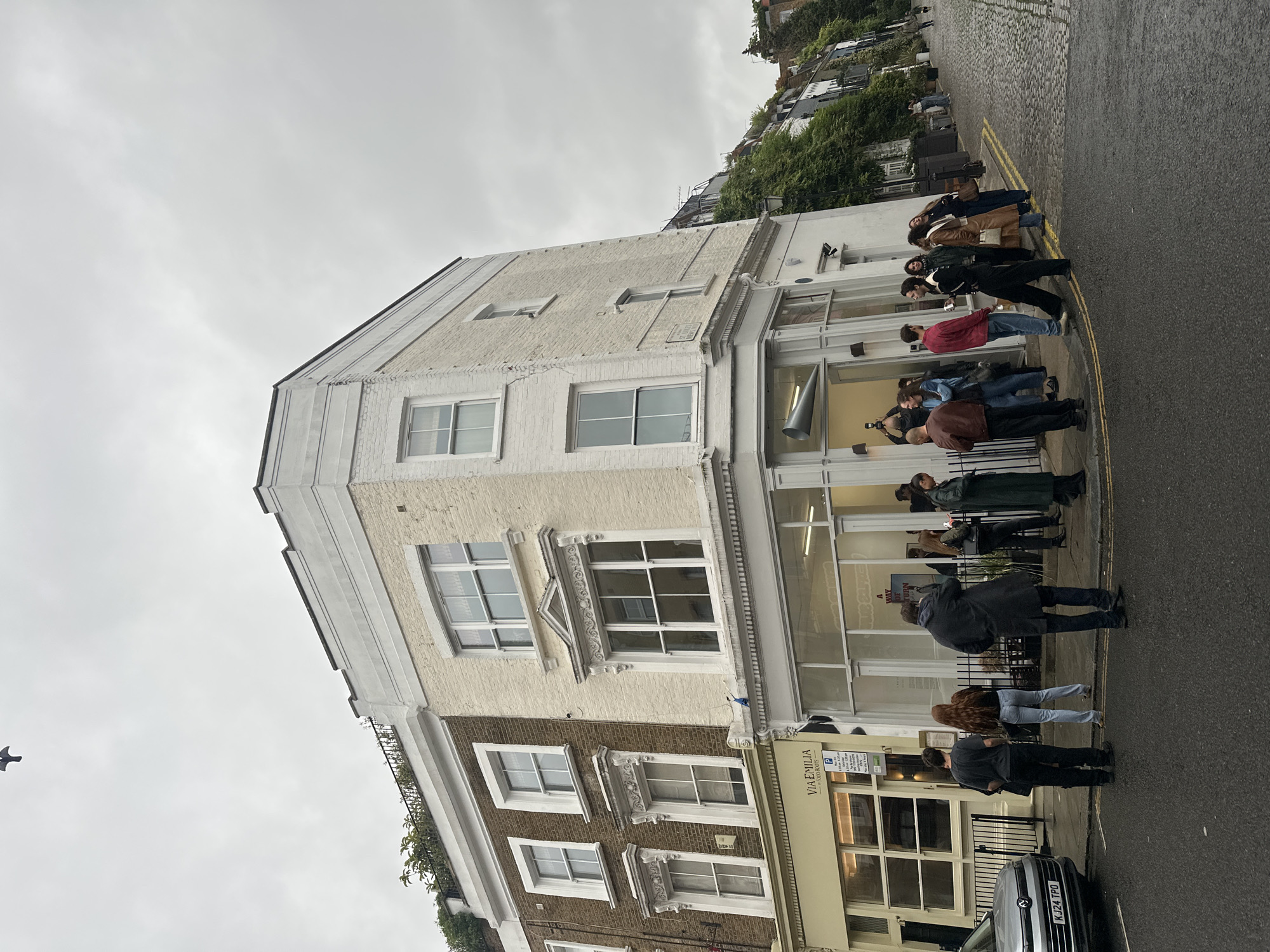The image depicts a street scene in London, United Kingdom, under an overcast sky. The primary focus is on the corner of a building with a cream-colored facade, housing a contemporary art gallery. A group of people, numbering approximately 15, are gathered outside the gallery, some looking at their phones and others engaged in conversation.
The building features large glass windows at street level, offering a glimpse into the gallery's interior, where a conical metal sculpture is visible. Above the gallery, the building has several windows on its upper floors. Adjacent to it is a brown brick building with similar architectural features.
In the foreground, a silver car is parked on the street, and a cobblestone pavement lines the street, curving around the corner. The environment suggests a commercial or artistic district within the city. The overall mood of the image is casual and observational.
