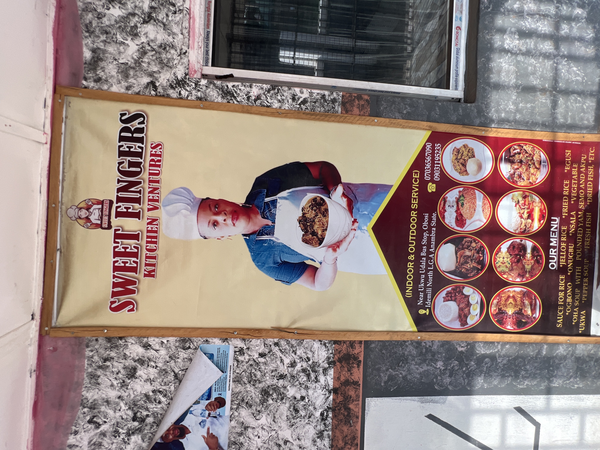 A close-up shot captures a vertical banner advertising "SWEET FINGERS KITCHEN VENTURES" in Nkpor, Nigeria. The banner is predominantly yellow and red, featuring a large image of a woman in a chef's hat and apron holding a bowl of food. Below her are several smaller images of various dishes. The text on the banner includes the restaurant's name, address, phone numbers, and a menu of offerings. The banner is mounted on a textured wall, possibly stucco or concrete, with some peeling wallpaper or paint in the background. The lighting suggests it's daytime, and the overall scene appears to be an outdoor or semi-outdoor setting, perhaps a storefront or market stall.