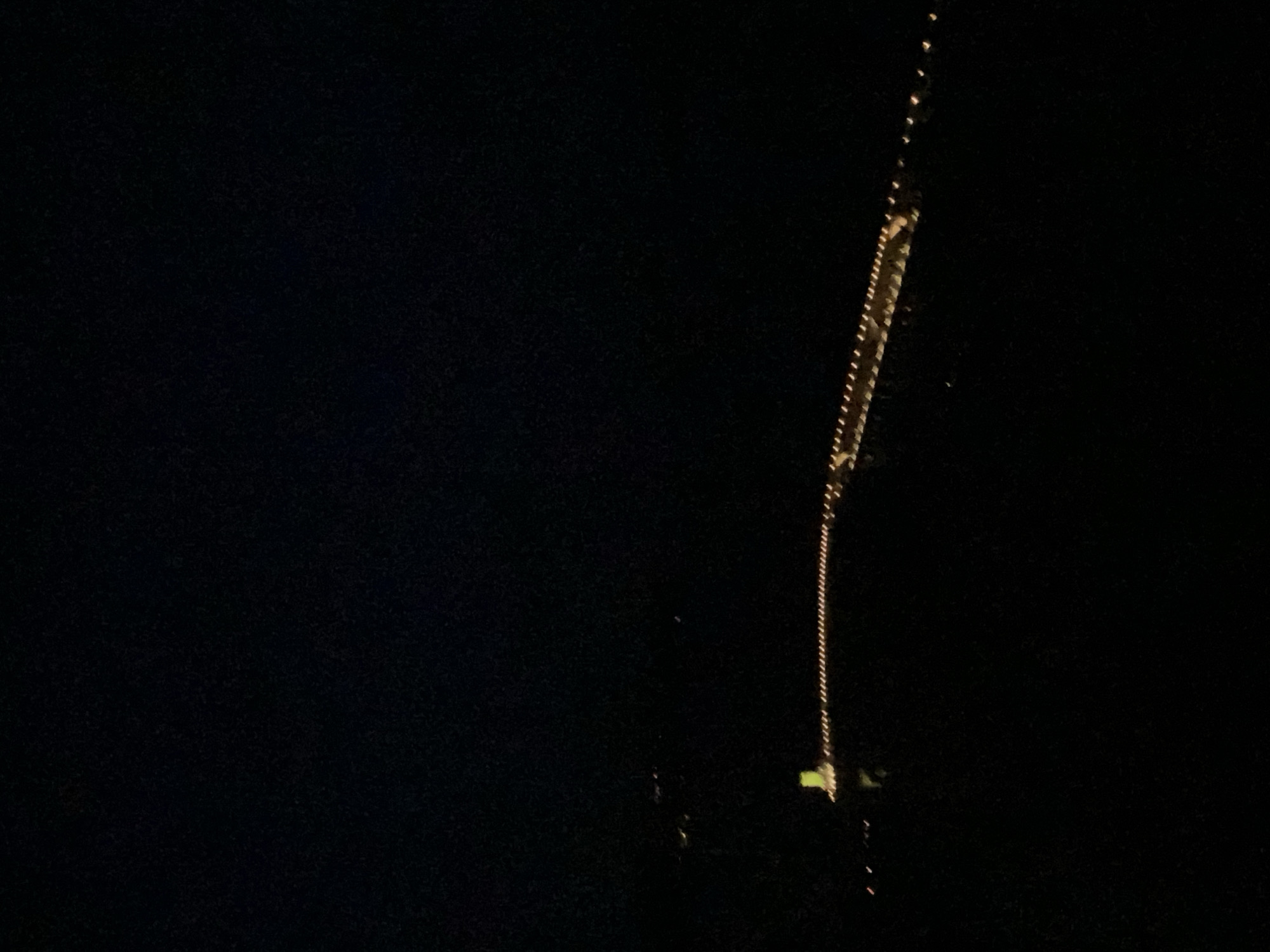 At night in Radovići, Montenegro, a bright vertical construction crane tower is decorated with closely spaced white lights. The crane is located on the right side of the frame and appears to be positioned against a dark background, suggesting the night sky or a shadowy area. The dark scene emphasizes the crane's illuminated form and its prominence in the surroundings.