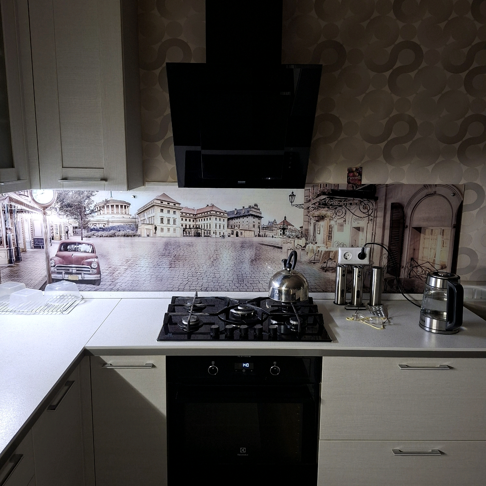 This is a forensic, matter-of-fact description of a domestic kitchen scene. The environment is a modern kitchen with light-colored cabinetry and a light-colored countertop. A large backsplash mural depicts an outdoor scene with cobblestone streets, historic European-style buildings, a vintage car, and a clock tower.
On the countertop, there is a stainless steel kettle, a black gas stovetop with four burners, and a black oven with a digital display showing "1:40". To the right of the stovetop, there are three stainless steel cylindrical objects, an electrical outlet with a plug, and a pair of eyeglasses. A glass kettle is also visible on the right side of the countertop.
Above the backsplash, a black, angled range hood is installed over the stovetop. The wall behind the range hood is covered in textured wallpaper with a repeating circular pattern. The cabinetry includes several drawers and doors with horizontal silver handles.
The image was taken in a kitchen located in Anapskaya, Russia. There are no people visible in the scene. The overall impression is one of a clean, well-equipped kitchen with a decorative backsplash.