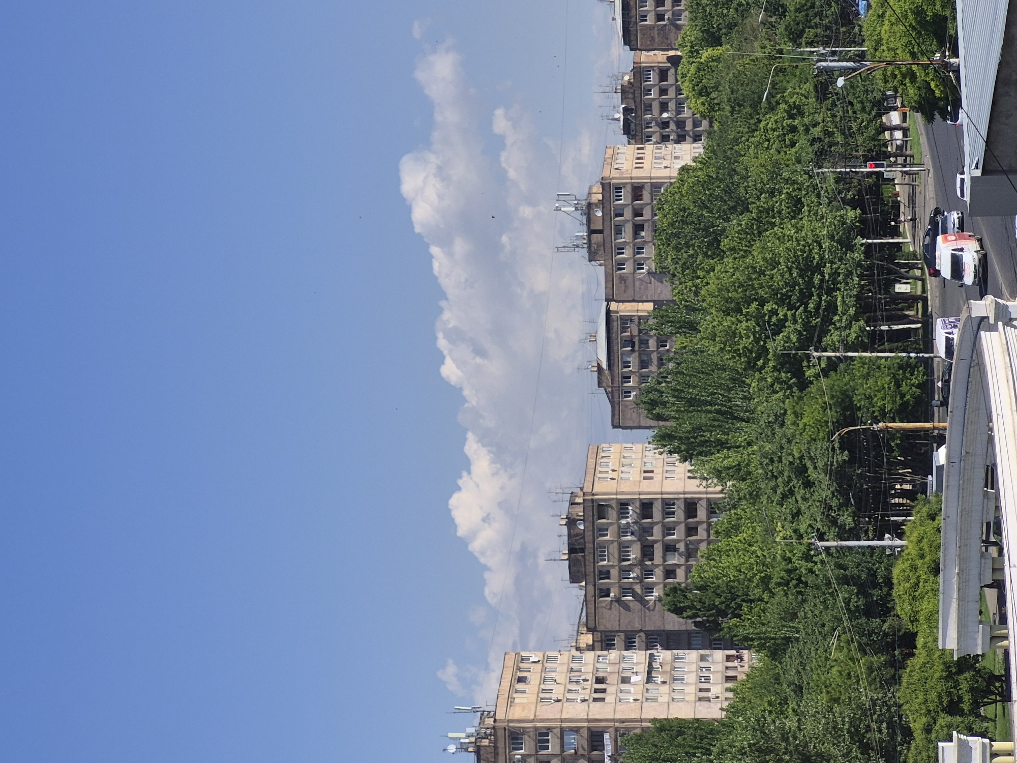 The image displays a sunlit urban landscape, as if viewed from an elevated position looking down a street lined with buildings and trees. The sky occupies the upper portion of the scene, a clear and bright blue, featuring a distinctive, elongated white cloud formation stretching horizontally across the middle. Several tiny dark specks, likely birds, are visible against the blue expanse.
Below the sky, a row of multi-story apartment buildings extends horizontally across the mid-ground. These structures exhibit a uniform, somewhat blocky architectural style, likely characteristic of Soviet-era construction, consistent with the Yerevan, Armenia context. They are colored in shades of beige and grey, with numerous rectangular windows, some of which have air conditioning units. Roofs show various antennae and satellite dishes.
A dense canopy of lush green trees forms a prominent line between the buildings and the street, spanning from the left to the right side of the frame. Through and beneath the trees, a paved street is visible. Several vehicles are present on the street, including a white van with red markings, and other cars further down. Utility poles, power lines, streetlights, and a red traffic light are visible elements of the urban infrastructure.
The overall scene depicts a typical bright, sunny day. The clear sky and strong illumination suggest it is mid-morning or afternoon. There is no visible text. The photograph captures a calm, everyday street view in a city setting.