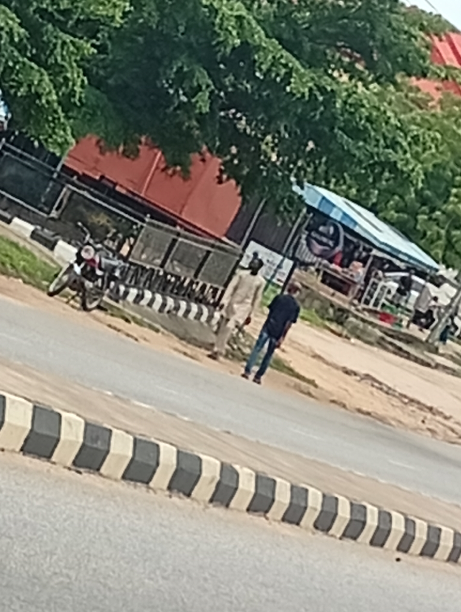 The image depicts a street scene in Adankolo, Nigeria, with a road in the foreground marked by black and white chevron barriers. To the left, a motorcycle is parked. Further back, two men are walking on the side of the road. Behind them, a market stall with a blue and white awning is visible, with several people browsing or working within. The background features lush green trees and a red-roofed building. The overall atmosphere suggests a typical day in a bustling Nigerian town.