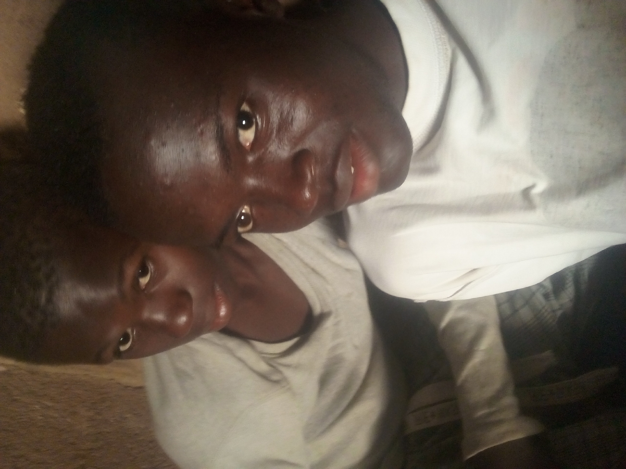 Two young Black individuals, likely male, are shown in a close-up, indoor portrait. They are positioned side-by-side, with the individual on the right closer to the camera and taking up more of the frame. Both individuals are wearing t-shirts, with the one on the left appearing to be a light grey or white, and the one on the right a bright white. The skin tones of both individuals are dark. The background is a textured, earthy brown wall. The lighting is dim, creating shadows and highlights on their faces, which appear slightly oily. The individual on the left is looking towards the camera with an open-mouthed expression, while the individual on the right is looking slightly to the left of the camera with a closed-mouth expression. Their faces are the primary focus of the image.