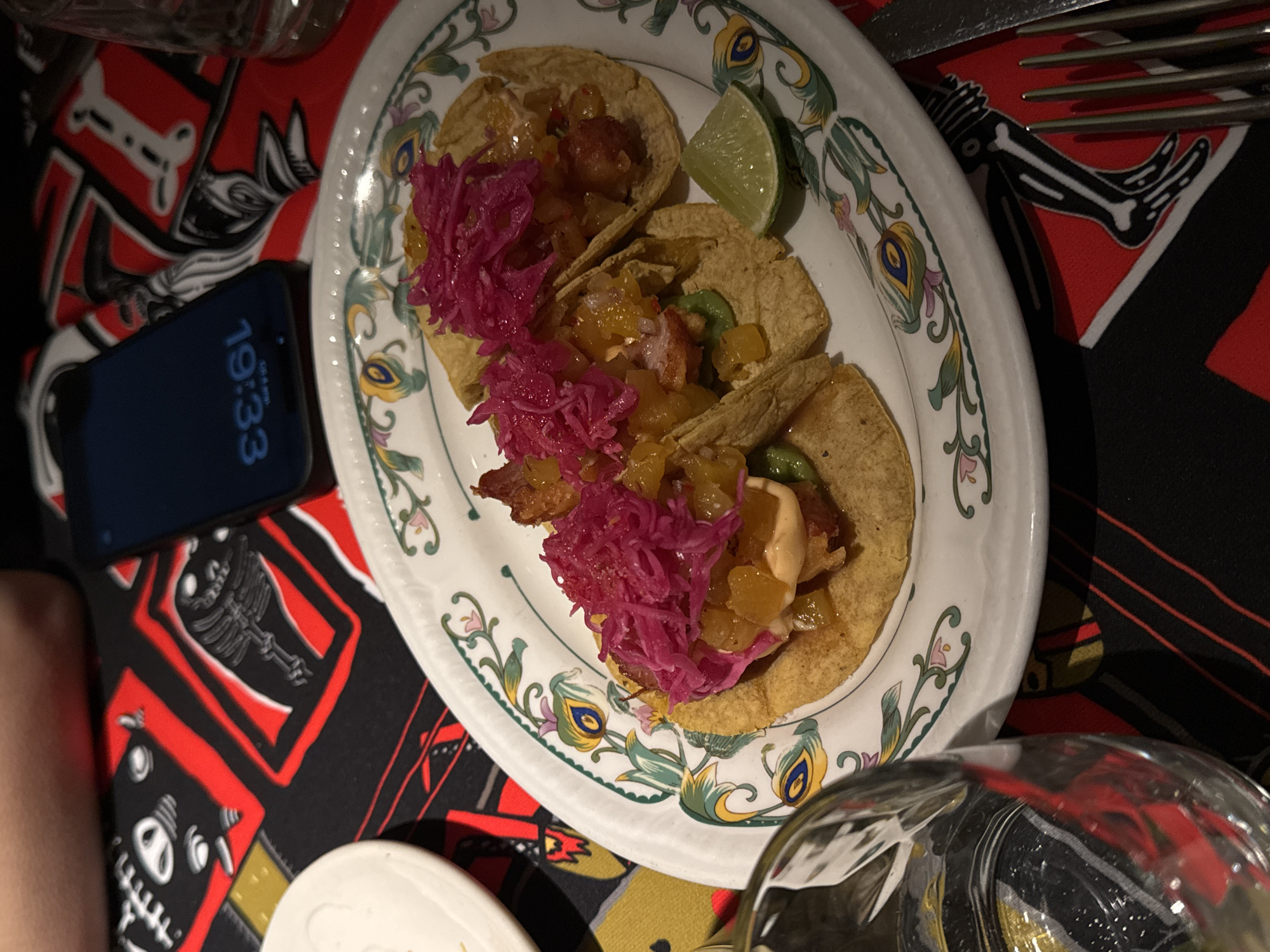 The image shows a plate of three tacos with red cabbage and a slice of lime on a patterned tablecloth in what appears to be a restaurant. A smartphone showing a time of 19:33 is positioned to the side of the plate. A partially visible glass of water is at the lower right. The tablecloth has a repeating pattern featuring stylized skeletons in red and black. There is no indication of this photo being in Moscow, Russia.