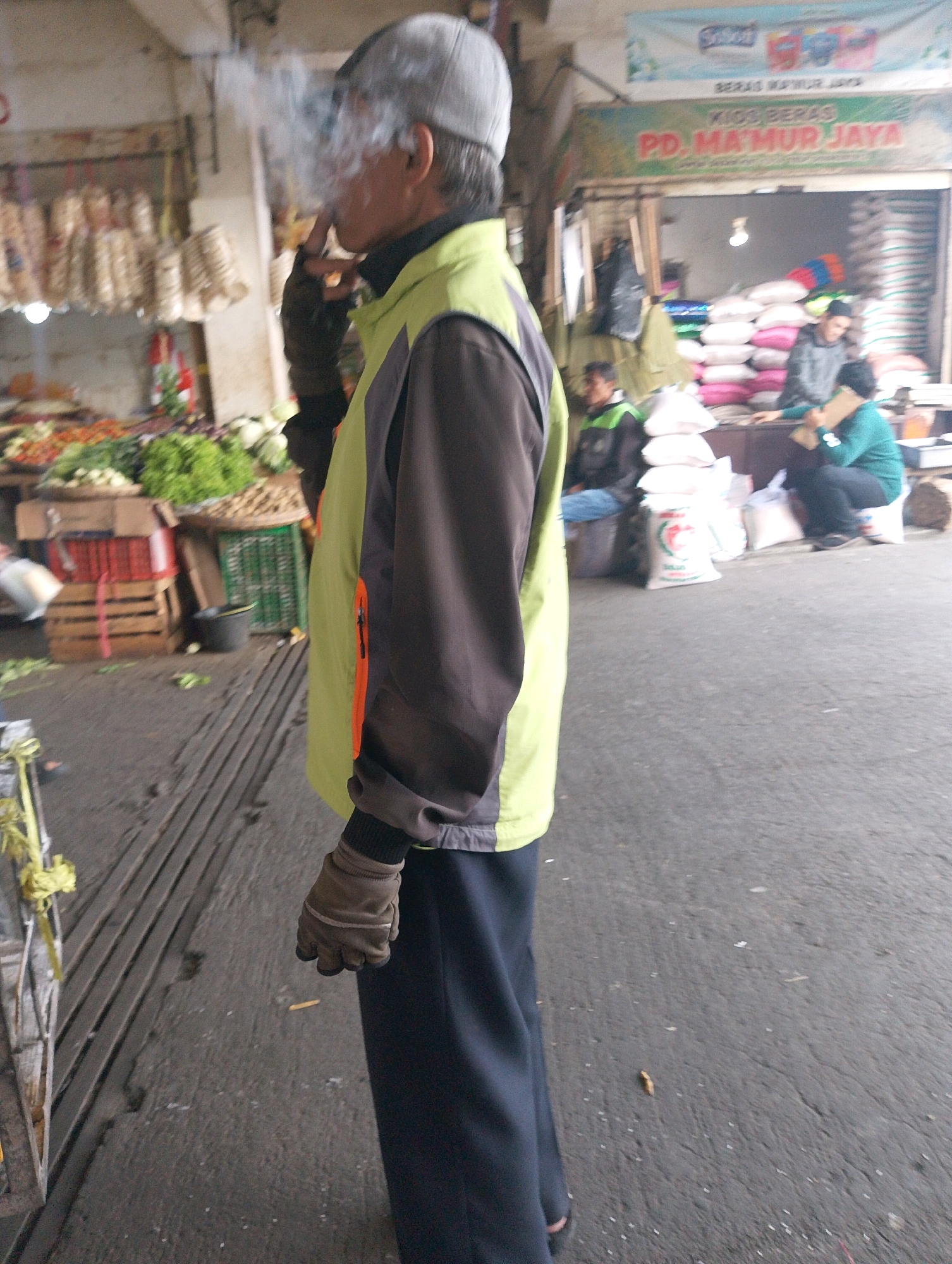 A person wearing a grey baseball cap, a green and brown jacket, and dark pants is standing and smoking. The person is wearing tan gloves and exhaling smoke. In the background, there is a market stall with various goods for sale, including produce and sacks of rice. Several other people are visible in the background, some appearing to be customers or vendors. The ground is paved with dark asphalt and some wooden planks. The overall scene suggests a busy market environment. The text on the stall's signage appears to be in Indonesian.