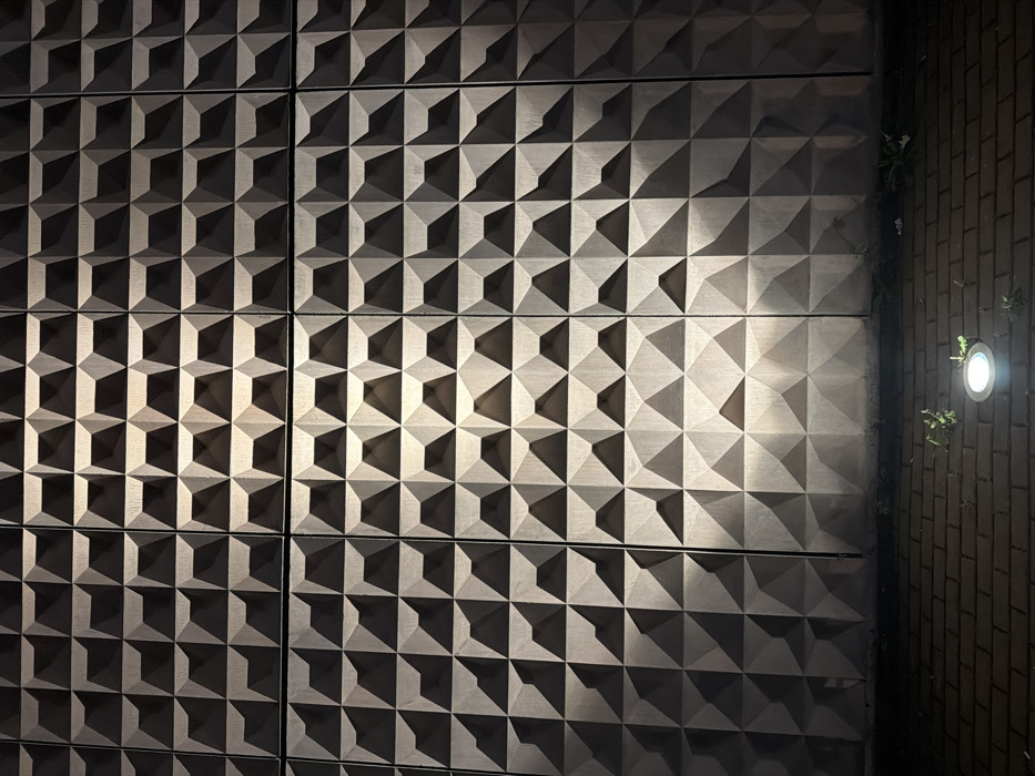 A nocturnal, low-angle photograph captures a segment of a building facade and an adjacent brick pavement in Penafiel, Portugal. The facade, constructed from large, rectangular panels of a grayish-brown concrete or stone-like material, displays a repetitive three-dimensional geometric pattern of inverted pyramidal indentations. The wall is unevenly illuminated, with the right side appearing brighter due to an unseen light source, and the pattern's relief emphasized by strong shadows and highlights. The ground consists of dark reddish-brown rectangular bricks arranged in a uniform pattern, serving as pavement. A single, circular, silver-rimmed LED uplighter is recessed into the brickwork in the lower-center, emitting a bright white light directly onto the wall. Sparse patches of small green vegetation are visible growing between the bricks, particularly around the light fixture. No individuals, animals, or animate subjects are present in the scene, and no specific actions or interactions are depicted.