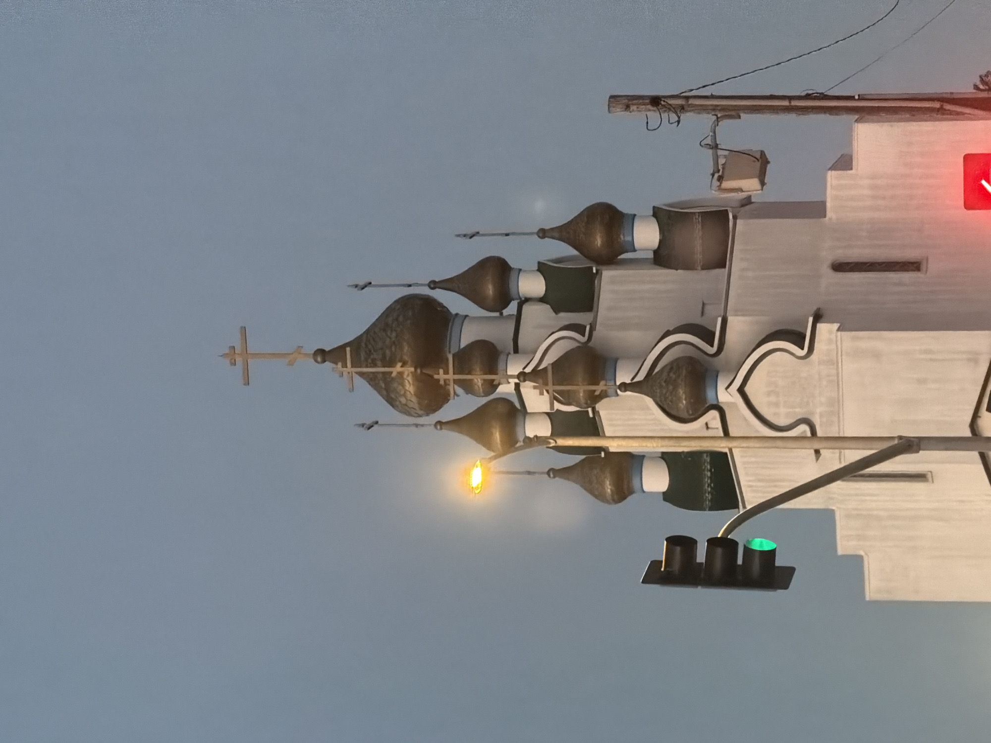 The image shows a close-up view of the domes and a cross atop an Eastern Orthodox church, possibly St. Nicholas Orthodox Church in Los Angeles, given the presence of a traffic light and utility pole consistent with urban California. The sky is overcast and pale blue, suggesting twilight or an overcast day. The church's white facade is visible, with several golden onion domes adorned with crosses. A bright yellow street light illuminates the scene, casting a glow on the church's architecture. In the foreground, a black traffic light shows a green signal, and a red signal is visible at the bottom of the frame. A wooden utility pole with wires stands to the right, with a small box-like device attached to an arm extending from it. The composition emphasizes the juxtaposition of religious architecture with modern urban infrastructure.