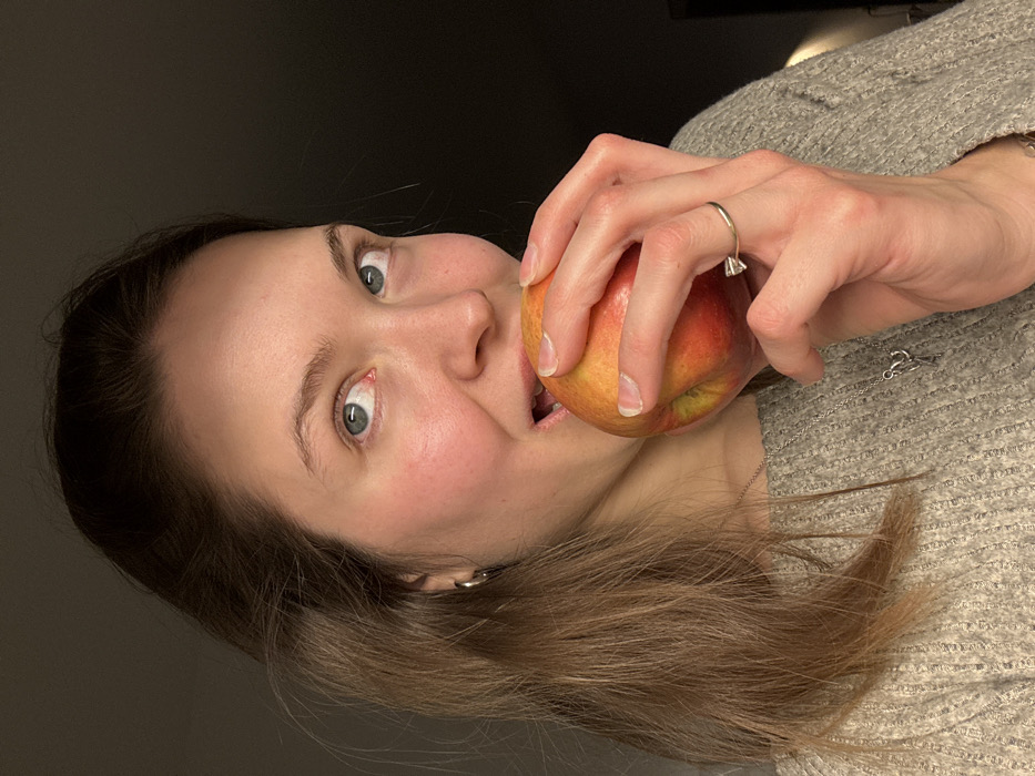 A light-skinned adult female is depicted in a close-up, chest-up portrait, holding a red and yellow apple to her mouth as if about to bite it. She has long, light brown hair, blue eyes, and her gaze is directed upwards and slightly to the right of the frame. Her mouth is open. She wears a light gray, possibly knitted, long-sleeved garment. A small silver-colored hoop earring is visible in her left ear, and a thin silver-colored chain with a small pendant hangs around her neck. On the ring finger of her left hand, which holds the apple, she wears a gold-colored ring with a single, prominent, clear stone. The background is uniformly dark, indicating an indoor setting with minimal visible environmental details. The precise location within the city of Tel-Aviv, Israel, cannot be determined from the visual information provided in the image.