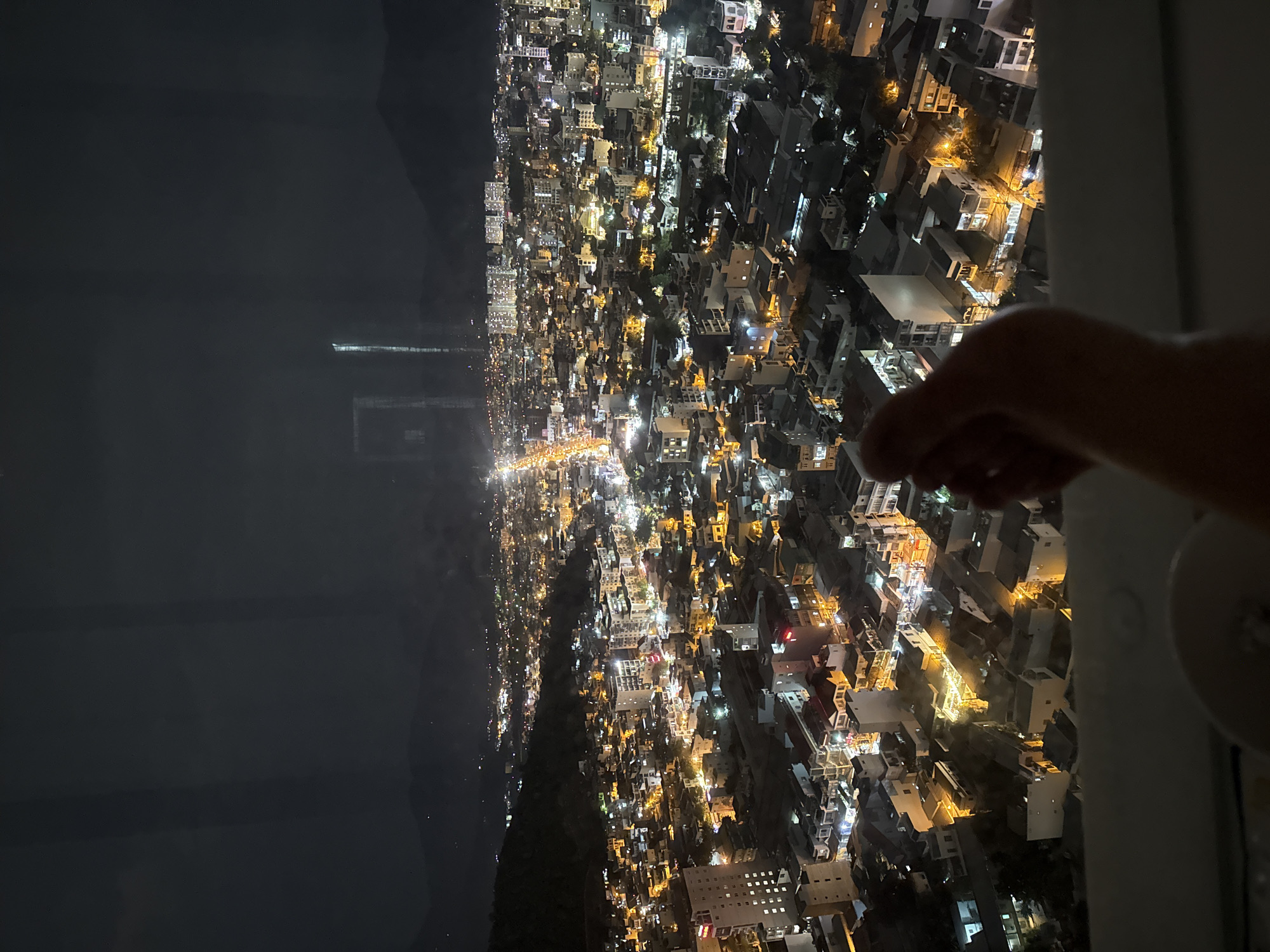 The image shows an aerial nighttime view of Nha Trang, Vietnam, characterized by a dense urban landscape illuminated by streetlights and building lights. The city sprawls across a plain and extends towards the base of dark, silhouetted mountains in the background. A prominent feature is a brightly lit street that cuts through the center of the city, suggesting a major thoroughfare. The architecture is predominantly low-rise, with many buildings appearing to be residential or commercial structures with visible windows lit from within. Some taller buildings are visible, particularly in the distance. In the foreground, a human hand is partially visible, positioned in a relaxed manner, with its shadow cast over the cityscape below. The hand appears to be resting on a surface, possibly a railing or window ledge, suggesting the vantage point is from a high-rise building or a balcony. The overall scene evokes a sense of urban activity and life continuing into the night.