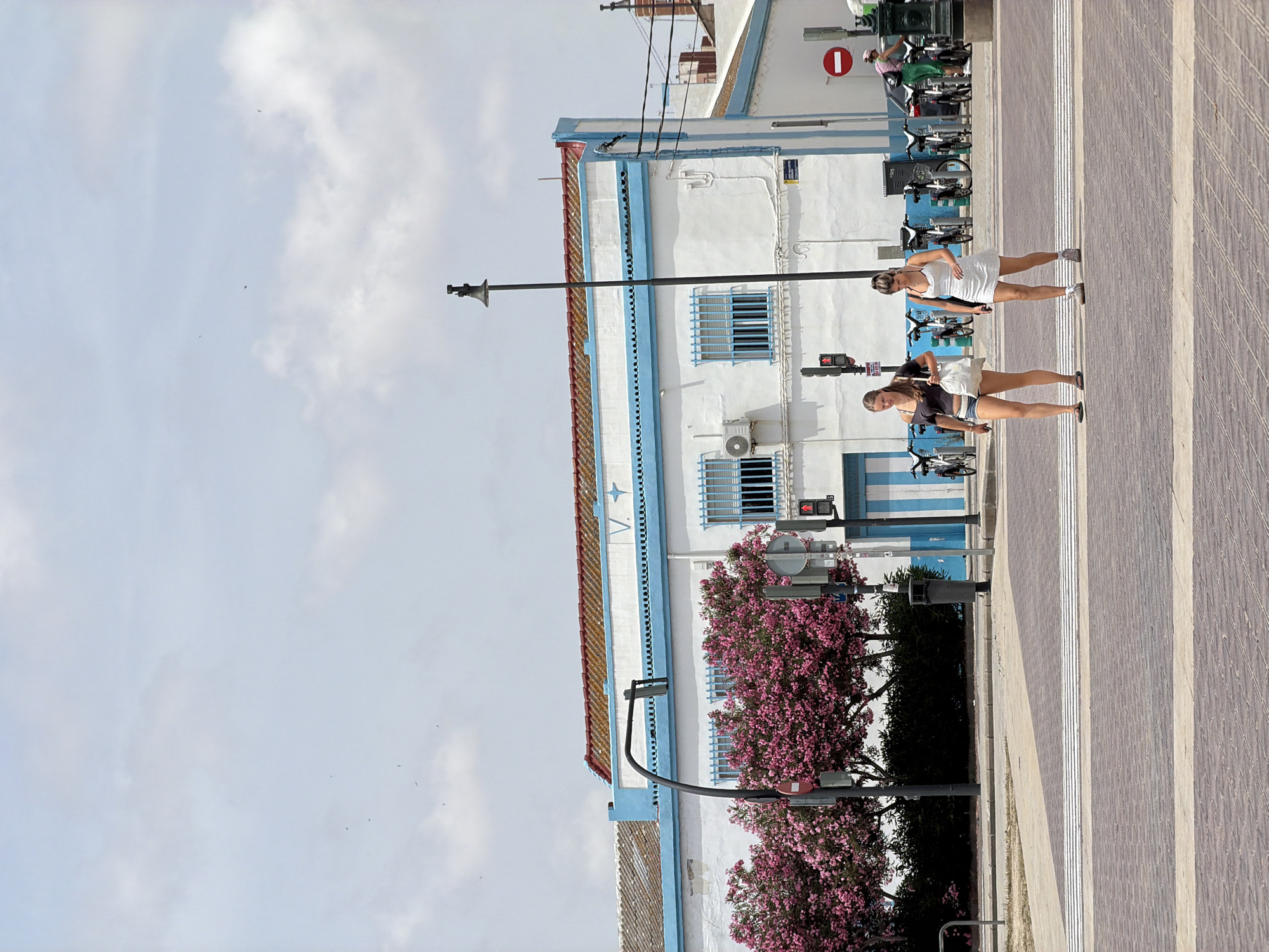 Two young women are walking across a pedestrian crossing in an urban environment. The woman on the left is wearing a dark off-the-shoulder top and denim shorts, carrying a white bag. The woman on the right is wearing a white sleeveless dress and sneakers. Behind them, a white building with blue trim and red-tiled roof is visible, with pink flowering trees in front of it. A lamppost and traffic signals are also present. The sky is overcast with some scattered clouds. In the background, a row of bicycles is parked.