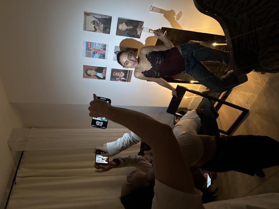An indoor scene in London, United Kingdom, depicts an impromptu photoshoot. In the foreground, left, a person with dark hair, wearing a white shirt, holds a smartphone horizontally, its screen displaying "9:49", aiming it towards the main subject. Immediately behind and slightly to the right, another person in a light long-sleeved top also holds a smartphone horizontally, aimed at the same subject, its screen showing an image.
The central subject, a female with dark hair pulled back, is positioned to the right of the frame. She wears a dark red camisole with black lace details and blue jeans. She holds a champagne flute in her left hand, which contains a clear liquid. Her right hand rests on a dark, reflective piece of furniture. She is looking towards the photographers. A pronounced shadow of her upper body is cast on the white wall behind her.
Affixed to the white wall are six posters arranged in two rows of three. These posters feature various portraits and scenes, including a child in academic attire, a woman in a pink dress with visible text "MON", two individuals in an interior setting, a woman with an American flag background, and another woman with dark hair.
The floor is wooden. Several tall, white taper candles are lit and positioned on the floor around the central subject and a dark, folding chair located center-left. An individual with a beard is partially visible, seated on the folding chair, observing the scene. A dark, ornate armchair is partially visible in the bottom right corner of the frame. Light-colored curtains are drawn on the left side of the room.