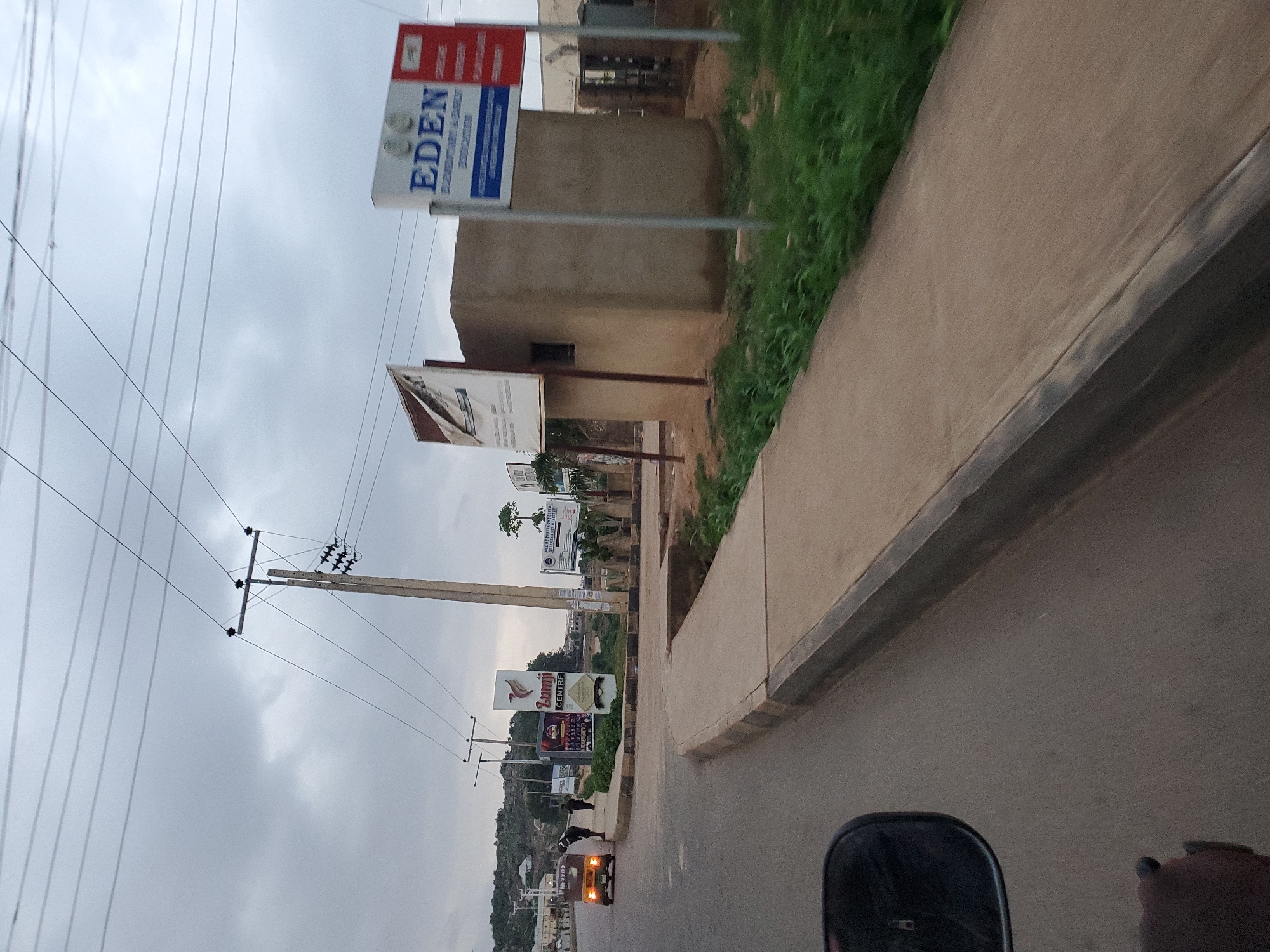 The image depicts a street scene in Gura Top, Nigeria, under an overcast sky. A beige-colored three-wheeled vehicle is parked on the right side of the road, facing away from the camera. Two individuals are standing near the vehicle. A paved sidewalk runs alongside the road, bordered by a concrete curb. Further down the road, a tall utility pole supports multiple overhead wires. A sign for "Zumji CENTRE" is visible, along with another sign displaying "EDEN ELEMENTARY & NURSERY EDUCATION". In the background, there are buildings and a densely vegetated hillside. The overall impression is that of a residential or commercial area in a Nigerian city.