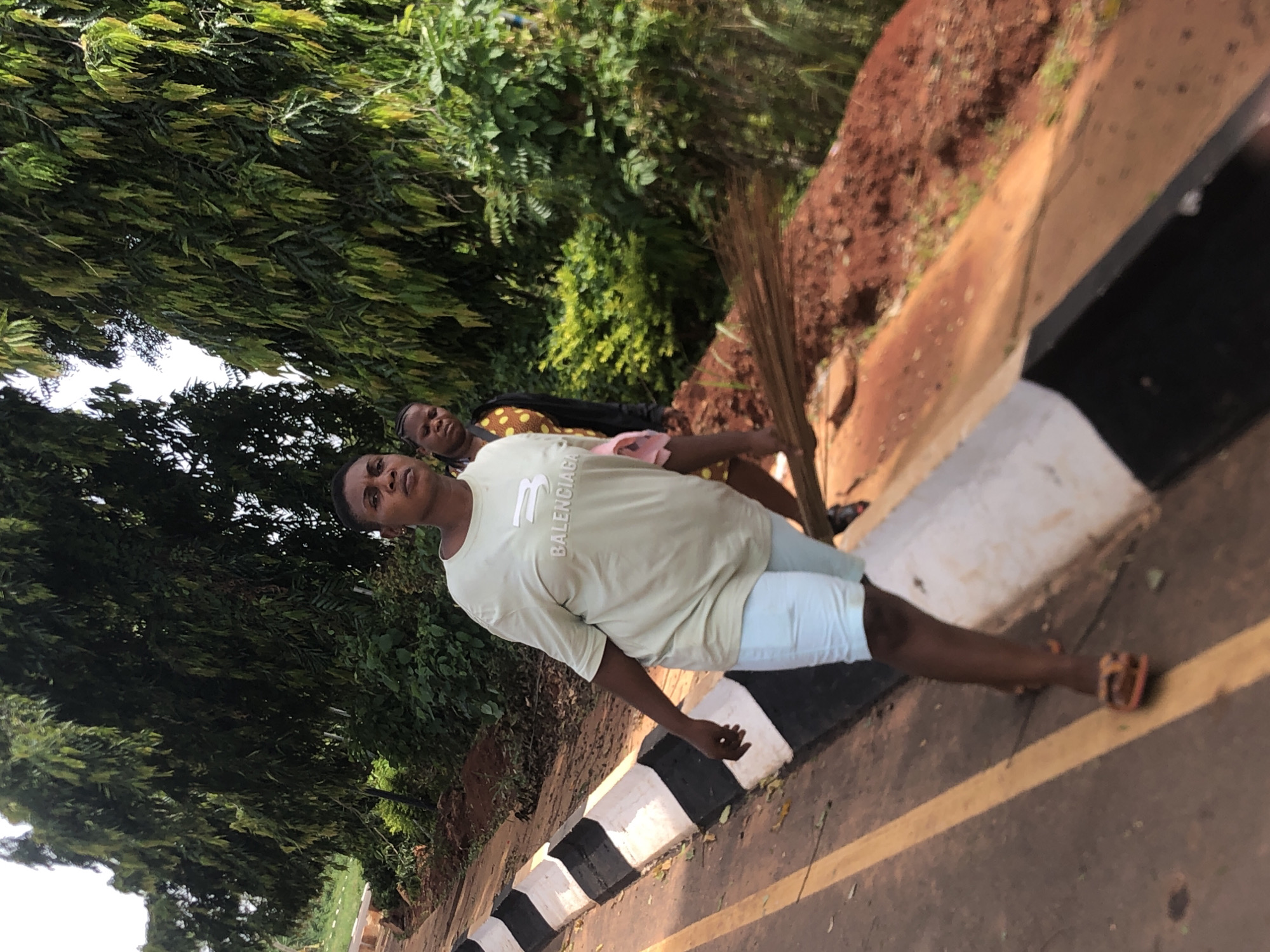 The image shows a man standing on a road in Nsugbe, Nigeria. He wears a light-colored t-shirt with "Balenciaga" on it, light blue shorts, and sandals. He stands near a black and white curb. In the background, another person is visible. Lush green foliage and reddish dirt embankment are present along the roadside.