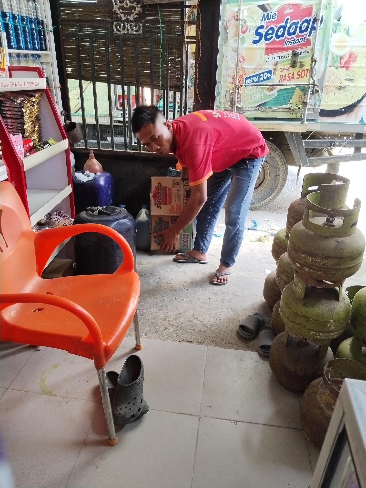 A man wearing a red t-shirt and blue jeans is in the process of unloading several boxes from a truck. The truck's side is decorated with a large advertisement for "Mie Sedaap Instant" with an image of noodles and various ingredients. In the foreground, there is an orange plastic chair. To the right of the man, a stack of green propane tanks is visible. In the background, a shelf unit with bottles of blue liquid and a display of packaged goods can be seen. The scene appears to be a small retail or roadside stall, possibly in Sawit Seberang, Indonesia, given the context. The ground is paved with light-colored tiles and concrete.