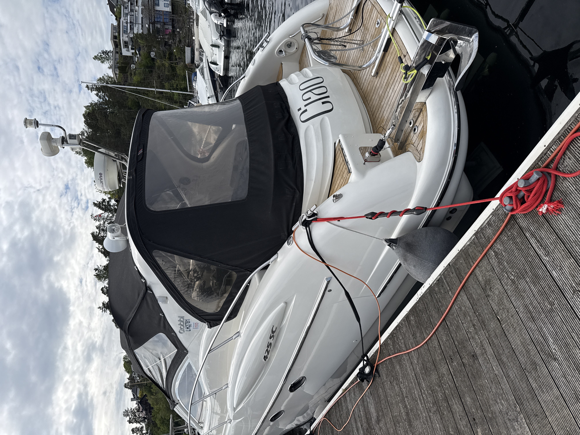 A partially-covered white boat is docked at a wooden pier. The boat is tied to the pier with a red rope. The name "CIRO" is visible on the boat's rear. "425 SC" is printed on the side of the boat, along with "Gobb LH2781." Gray fenders are between the boat and the pier. Other boats are visible in the background, moored in the water. Buildings and trees are visible on a hill in the background. The photo was taken in Nesøya, Norway. The sky is cloudy.