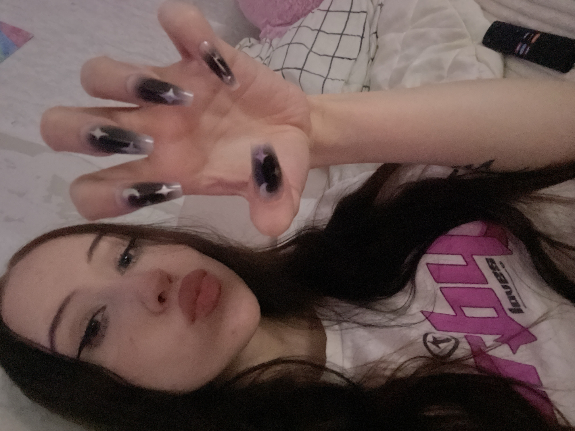 A young woman with long, dark hair is shown from the chest up, her right hand extended towards the camera. She is wearing a white t-shirt with a pink graphic design on the front. Her nails are long and clear with black tips and white star and moon designs. She has winged eyeliner and is puckering her lips. The background appears to be a bed with white linens and a pillow with a black and white checkered pattern. A black remote control is visible in the lower right corner. The scene is indoors, with soft lighting. There is no indication of the location within the city of Votkinsk, Russia.