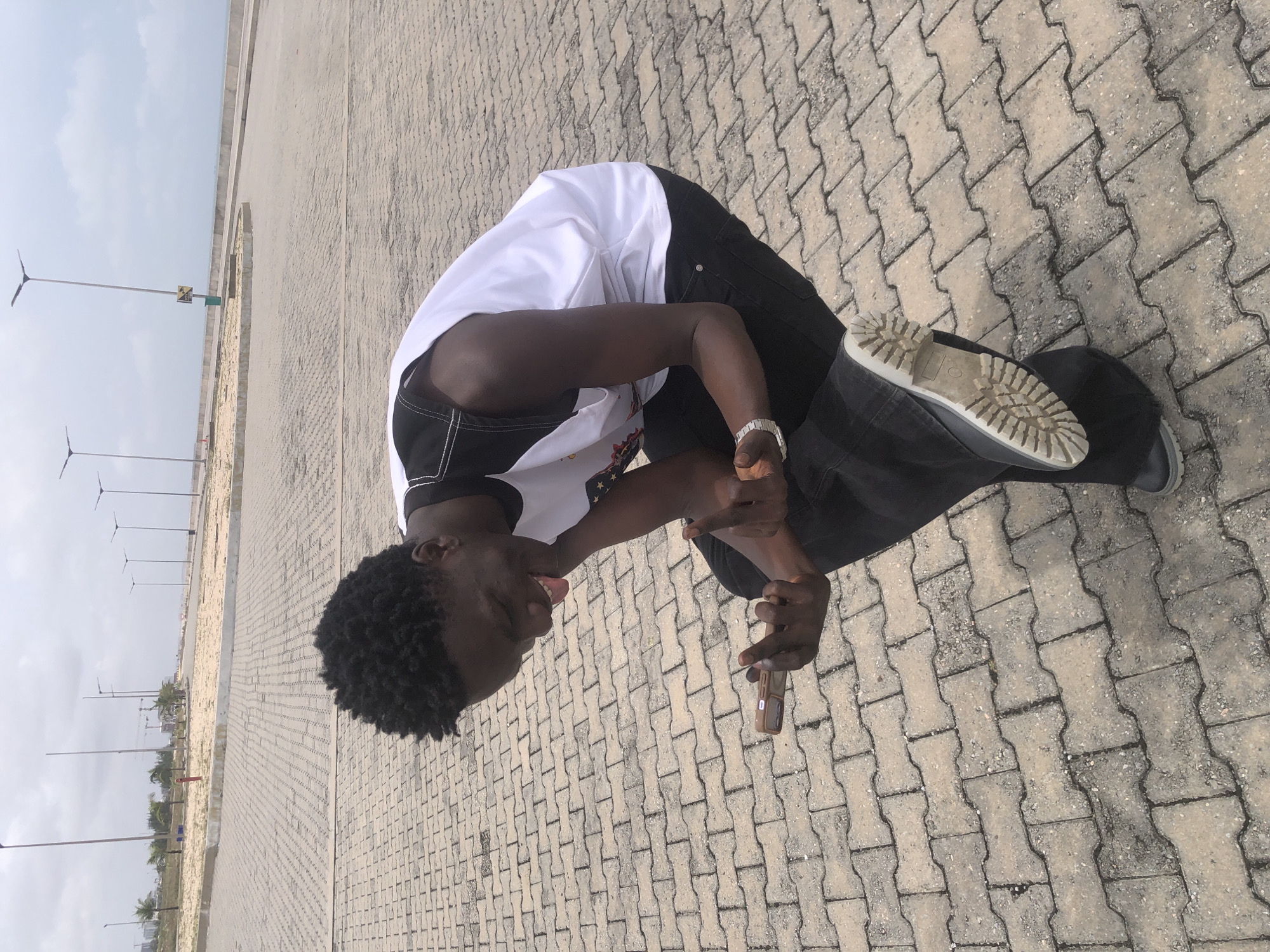 A Black man wearing a white and black sleeveless shirt and black pants is crouching outdoors on a paved walkway. He is holding a smartphone in his left hand and his right hand is raised, with his middle finger extended. His head is tilted back and he is laughing with his tongue sticking out. The background shows a paved area with streetlights and a body of water in the distance. The location appears to be a public outdoor space in Ikoyi, Nigeria.