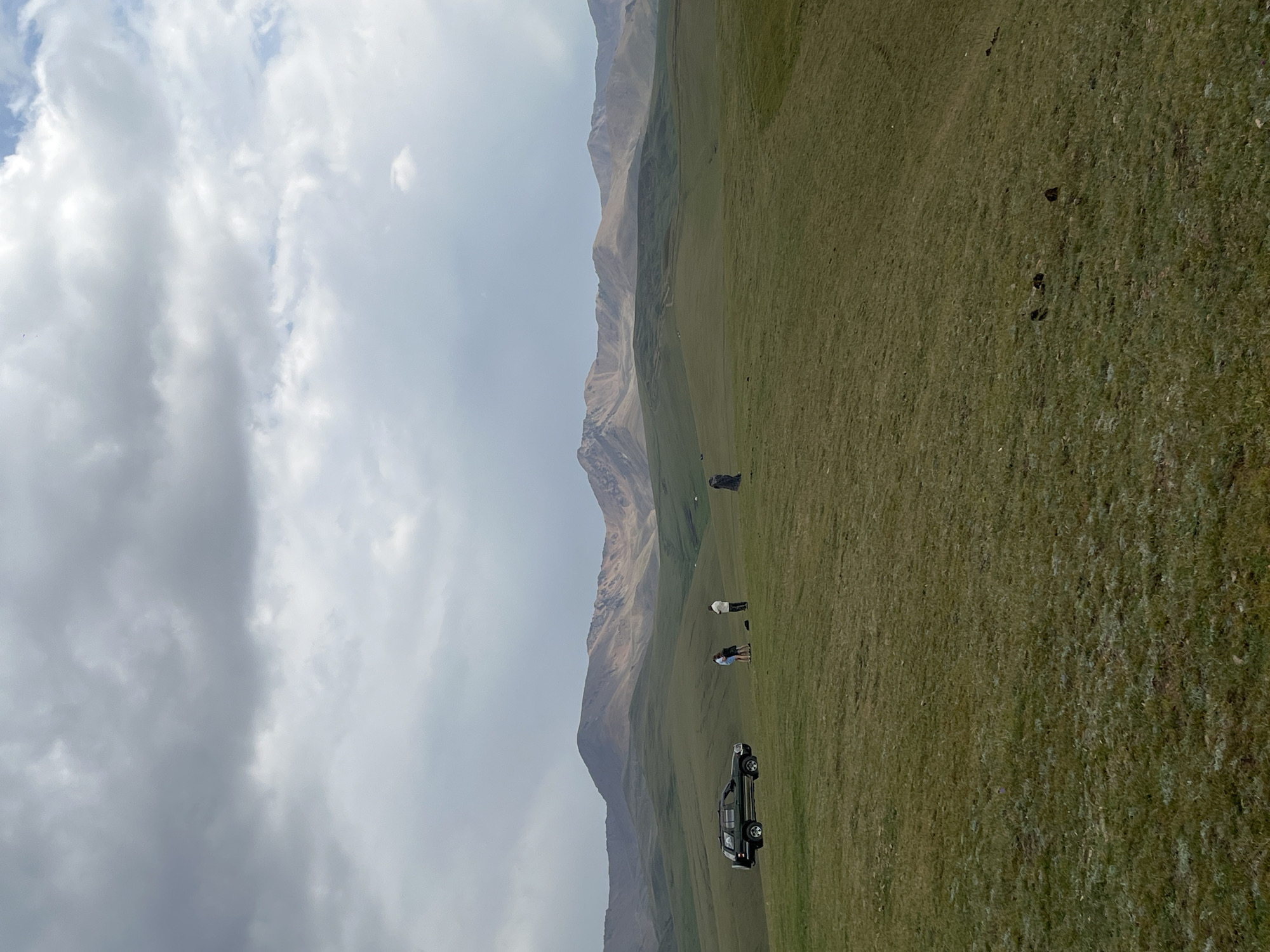 The image depicts a vast, grassy plain with rolling hills leading to a majestic mountain range in the distance. The sky is overcast with dramatic clouds. In the middle ground, a dark-colored SUV is parked on the left side of the frame. Near the SUV, three individuals are standing on the grass. One person, wearing a dark outfit, is standing a distance away to the right of the SUV and the other two individuals. The terrain is covered in short, green grass with some patches of lighter vegetation and scattered dark objects that appear to be animal droppings. The overall scene is natural and serene, suggesting an outdoor excursion in a remote, mountainous region. The specific location within the city of Batan, Kazakhstan, cannot be determined from the visual information provided.