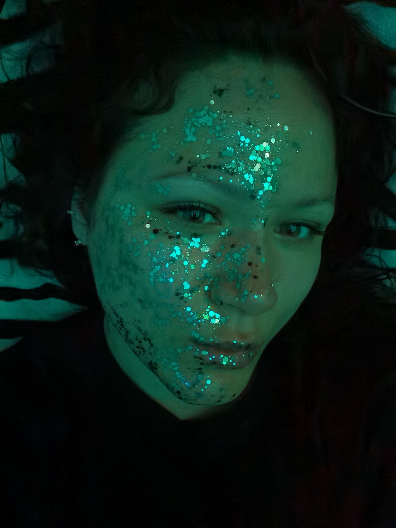 A close-up, high-angle portrait features a young woman with dark, curly hair, lying down and looking directly at the camera. Her face is extensively adorned with shimmering, multi-sized glitter predominantly in shades of teal and blue-green, catching the light. The glitter is concentrated on her forehead, cheeks, nose, and chin, with some smaller, darker specks intermingled.
Her skin appears to be illuminated by a strong green or teal light source, casting a vibrant hue across her face and highlighting the reflective glitter. Her eyes are open, with visible eyelashes and dark pupils, and she maintains a neutral to slightly contemplative expression. Her dark hair frames her face, appearing spread out against a very dark, indistinct background, suggesting a dimly lit indoor environment. She is wearing what appears to be a dark top. No specific location cues for Chaikovsky, Russia, are visible in the image itself, beyond the provided context. There is no visible text. The overall atmosphere is artistic and stylized, potentially related to a party, festival, or creative photoshoot.