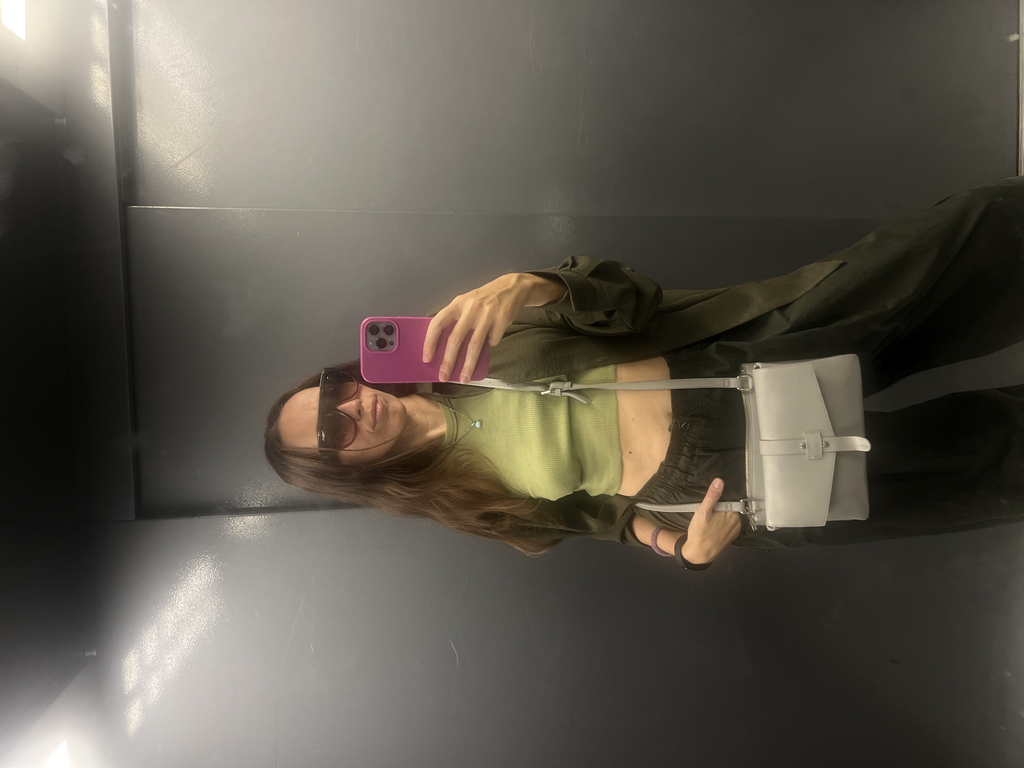 A young woman is taking a selfie in what appears to be an elevator. She is wearing a light green crop top, an olive green jacket, and dark pants. She is holding a pink phone in her right hand. A grey purse hangs from her shoulder. She is wearing sunglasses. The background is a grey metal elevator wall. The location cannot be determined as Moscow, Russia from the image.