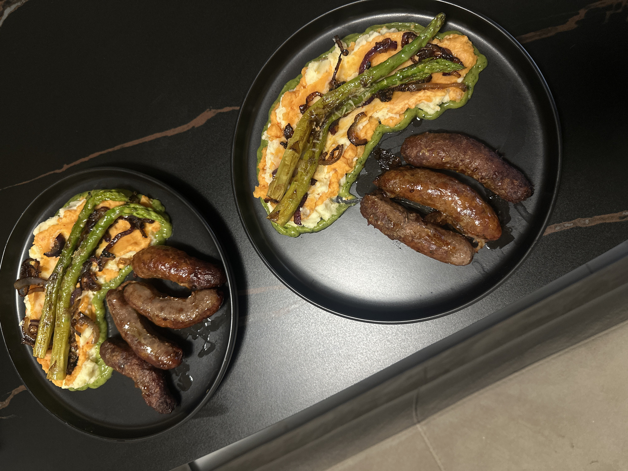 Two black plates of food are positioned on a dark, possibly granite, countertop with a light brown vein pattern. The plates contain what appears to be a combination of mashed potatoes, sweet potatoes, asparagus, caramelized onions, and sausages. Each plate holds a similar composition of these food items. The countertop is partially illuminated, and a section of a vertical surface, potentially a cabinet or wall, is visible in the lower right corner of the image. This scene gives no indication of the location being Sofia, Bulgaria.
