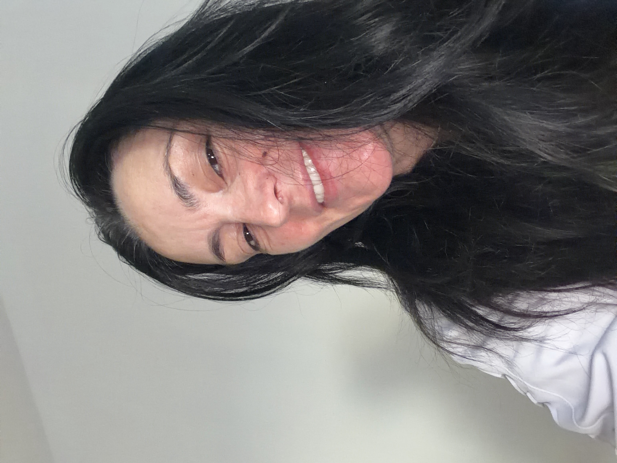 A close-up, low-angle, selfie-style shot shows a woman with long, dark hair and fair skin. Her head is tilted, and she is looking towards the camera with a slight smile, revealing her teeth. She is wearing a white top. The background is a plain, light-colored wall. The image was taken in Marl, Germany.