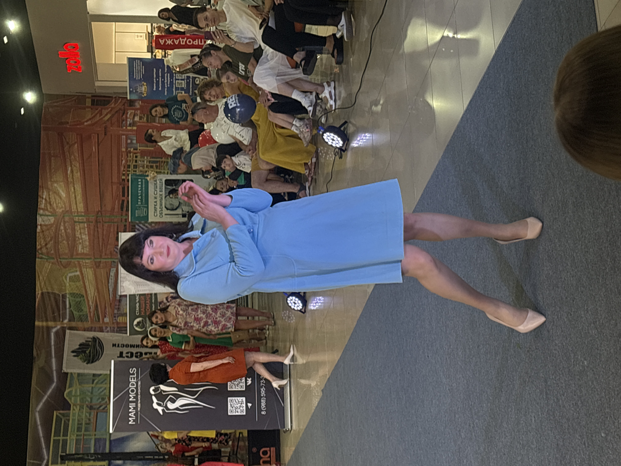 The image depicts a fashion show at a shopping mall, likely in Tri Potoka, Russia, based on the context of the provided crops and typical mall layouts in Russian cities. A woman in a light blue dress and high heels is the central subject, standing on a blue carpeted runway. She is looking towards the camera and appears to be posing or walking.
In the background, several other individuals are seated, observing the event. Some are dressed casually, while others are in more formal attire, suggesting a diverse audience. Various banners and signs are visible, indicating sponsorships or event information. One prominent banner reads "MAMI MODELS," suggesting this is a modeling event. Another sign in red letters above a store entrance says "zola." Lighting equipment, including spotlights, is positioned on the floor, illuminating the runway. The environment appears to be an indoor shopping mall with a tiled floor reflecting the lights and the surrounding activity. The overall scene conveys a lively atmosphere of a public event.