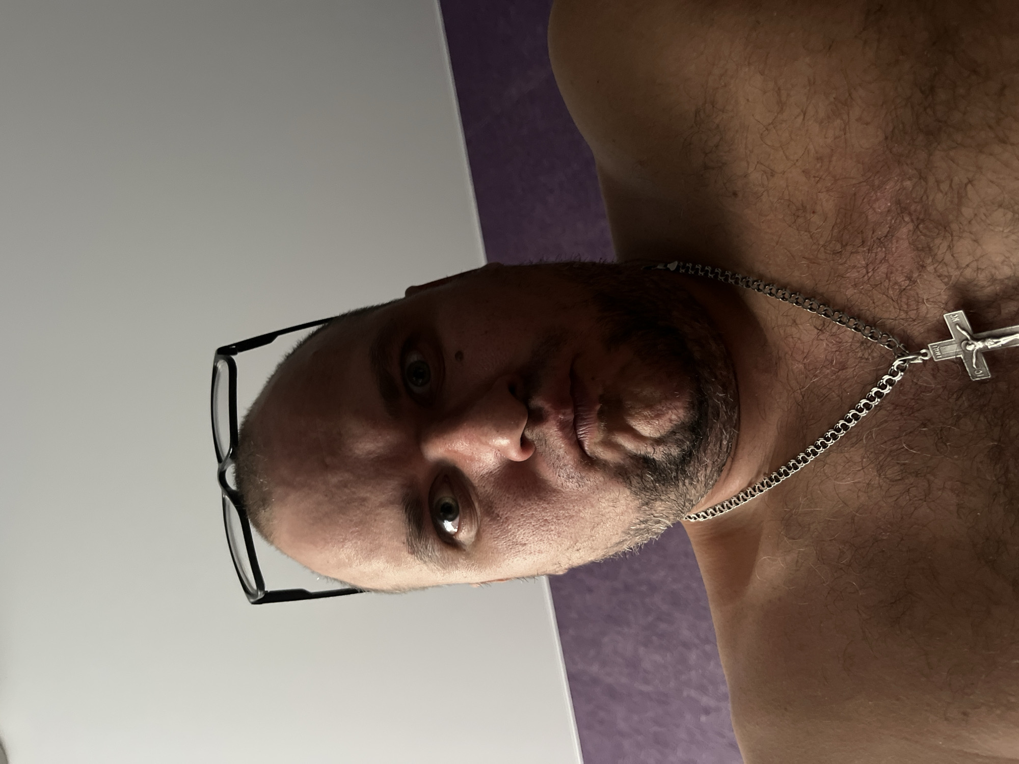 A low-angle, close-up, selfie-style shot captures a shirtless person with glasses perched on their forehead, looking directly into the camera with a pouting expression. The individual has a shaved head, a beard, and is wearing a silver chain with a cross pendant. The background consists of a light-colored wall and a purple textured wall. No specific location in Minsk, Belarus can be identified from the image.