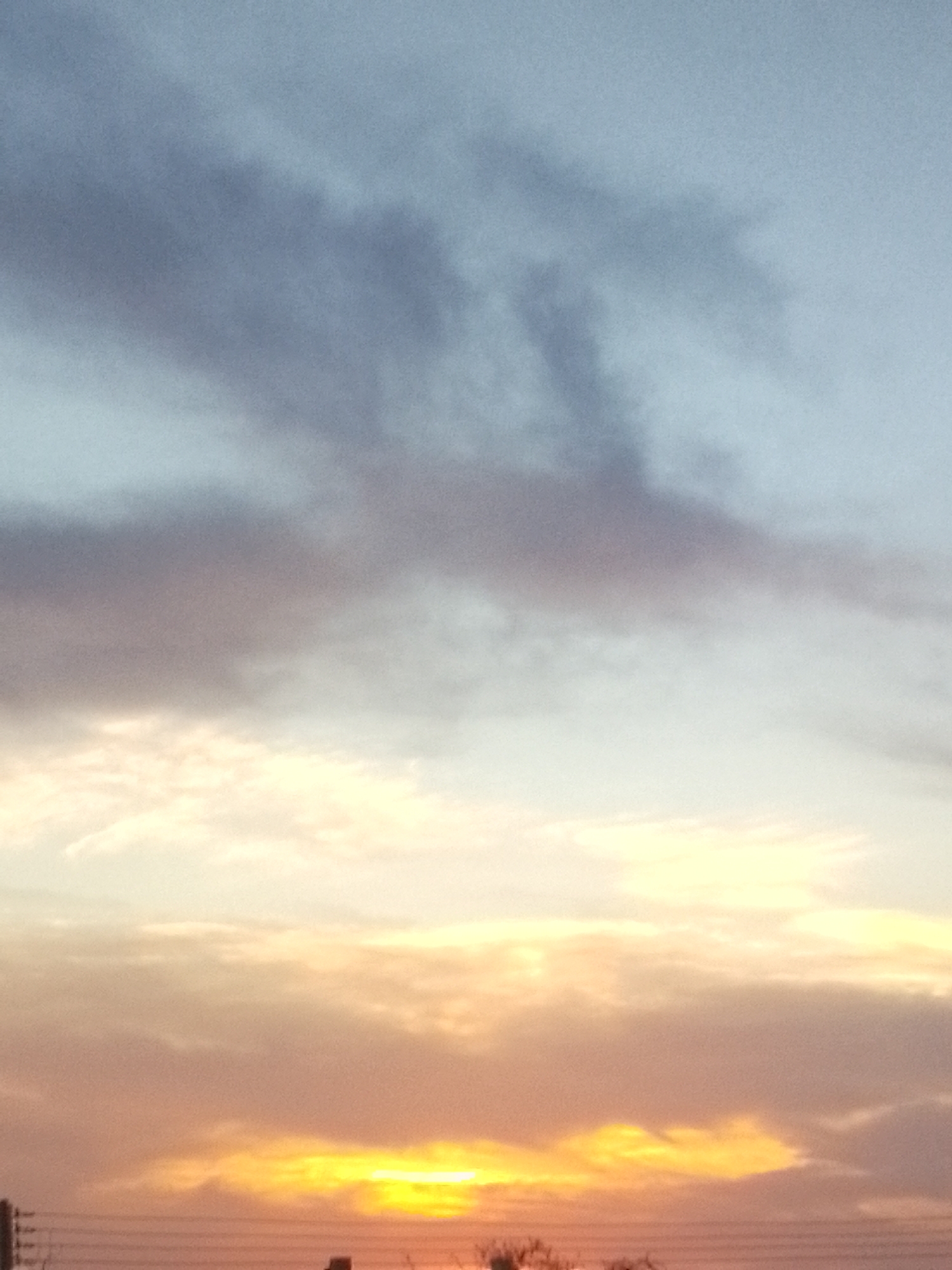 This media file captures a dramatic sky scene at either dawn or dusk, set over Juan Griego, Venezuela. The dominant subjects are the expansive sky and various cloud formations, illuminated by the low-lying sun.
The upper portion of the sky is a soft, pale blue, gradually transitioning downwards. Mid-frame, darker, denser clouds appear in shades of muted grey and lavender, showing subtle textures and forms. Below these, the sky brightens considerably, displaying a gradient of light cream, soft white, and warm orange tones.
At the very bottom of the image, the sun is partially obscured by clouds, appearing as a brilliant, intense band of yellow-orange light stretching across the horizon. This radiant glow bathes the surrounding clouds in warm, vivid hues, indicating a powerful light source characteristic of either sunrise or sunset. Given the context of Juan Griego, a sunset over the Caribbean is a strong possibility.
Faint, dark linear elements, possibly power lines or wires, are visible stretching horizontally across the lowest part of the frame. Below these lines, indistinct dark shapes suggest distant structures or vegetation, subtly hinting at a terrestrial foreground without providing clear identifying features.
There are no people or specific man-made objects clearly identifiable, nor is there any visible text. The image focuses entirely on the natural beauty and atmospheric effects of twilight, characterized by the striking interplay of light and color across the clouds.