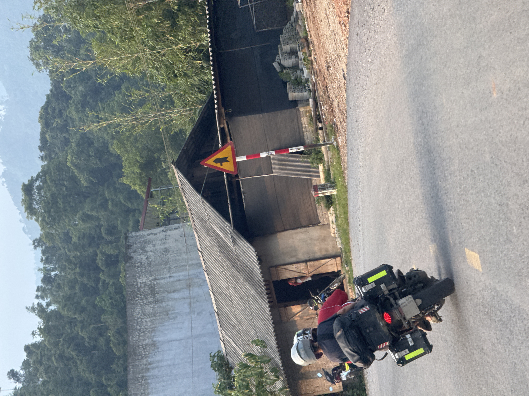 This image depicts a rural scene in Tân Quang Commune, Vietnam, bathed in natural daylight, likely morning or late afternoon. In the foreground, an adventure touring motorcycle is positioned on an asphalt road, facing away from the viewer. The motorcycle is heavily equipped with black panniers and a top case, and a chainsaw is notably strapped to its rear. A person wearing a white helmet is seated on the motorcycle.
To the left of the road, a simple building with a corrugated roof stands, partially obscured by lush green foliage. A concrete wall forms part of the structure, and stacks of concrete blocks are visible nearby. A yellow triangular road sign with a black arrow pointing to the left is mounted on a red and white striped pole, situated between the building and the road. Another person, wearing a red top, is partially visible in the doorway of the building.
The background is dominated by dense, forested mountains under a clear, bright sky, indicative of Vietnam's natural landscape. The scene conveys a sense of travel through a mountainous, rural area. There is no discernible text on any objects.