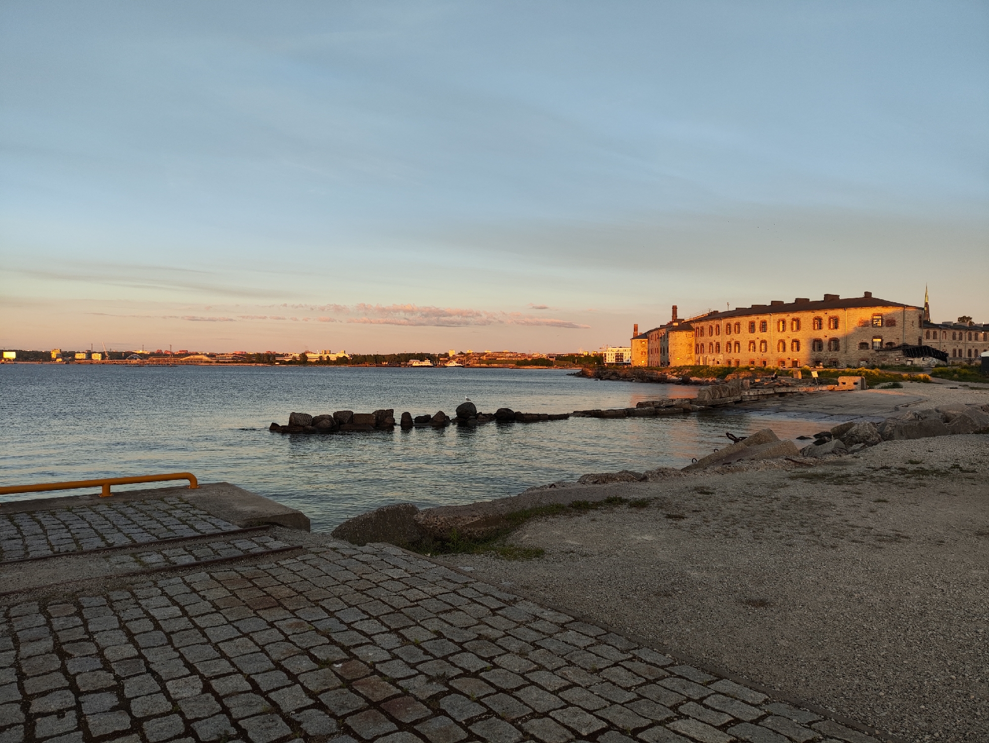 The image depicts a waterfront scene in Tallinn, Estonia, during what appears to be sunset. In the foreground, a paved area with cobblestones leads towards the water. Adjacent to this, a yellow railing is visible on a concrete structure. The water is calm, with a few rocks jutting out from it, some forming a small breakwater. On the right side of the image, a large, historic-looking building with multiple windows and chimneys is illuminated by the warm glow of the setting sun. The building has a long, rectangular shape and appears to be constructed of stone. In the background, the silhouette of a city skyline is visible across the water, suggesting an urban setting. The sky is a pale blue with wispy clouds, and the overall atmosphere is peaceful.
