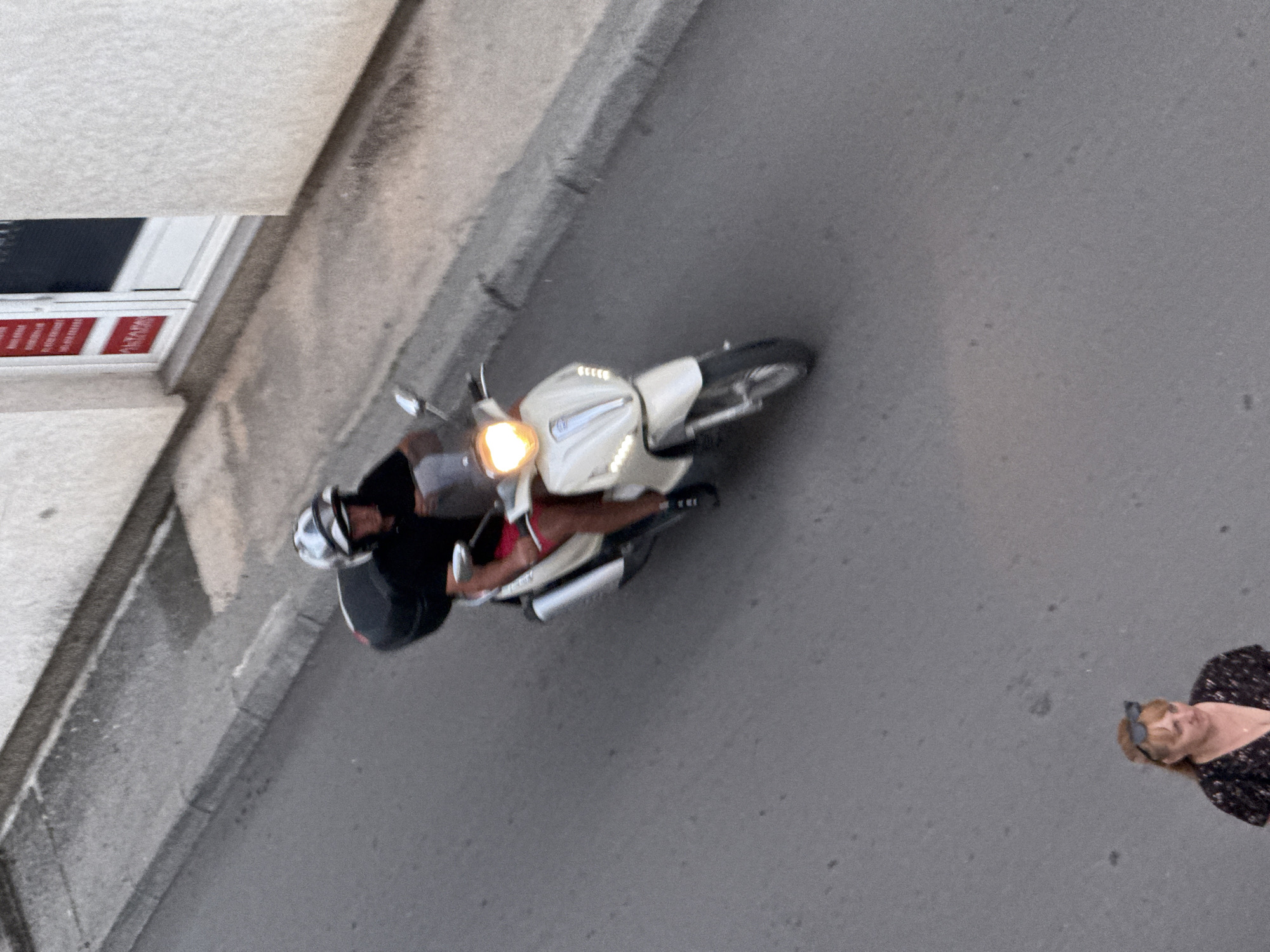 In Budva, Montenegro, a person wearing a black t-shirt, red shorts, and a white and grey helmet is riding a white scooter. The scooter has a black storage box on the back and its headlight is illuminated. The rider is facing away from the camera. In the foreground, a woman with blonde hair and sunglasses perched on her head is visible, wearing a dark floral-patterned top. They are both on a paved road next to a grey concrete curb. A building with a white textured facade and a white-framed door with red signage is to the right. The scene appears to be an outdoor street setting.