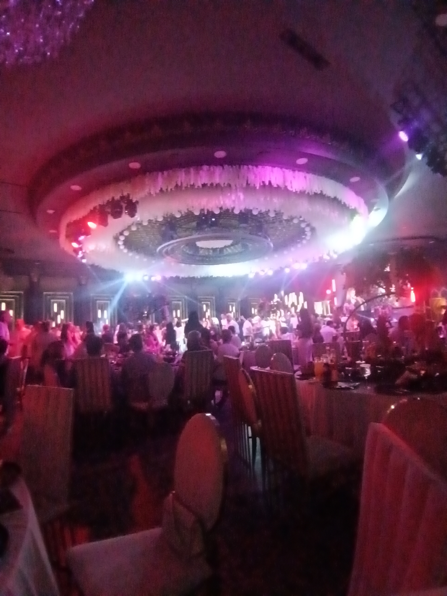 The image depicts an indoor event space, likely a banquet hall or restaurant, filled with people seated at tables. The ceiling features elaborate decorative lighting and a circular ceiling fixture. The ambiance is illuminated by colorful stage lights, predominantly in shades of pink and purple, casting a dramatic glow on the scene. Several individuals are visible in the background, some appearing to be standing or mingling. Tables are covered with tablecloths, and some have drinks and other items on them. The overall impression is of a lively gathering, possibly a celebration or a formal dinner. The image's context suggests it was taken in the city of Vagharshapat, Armenia.