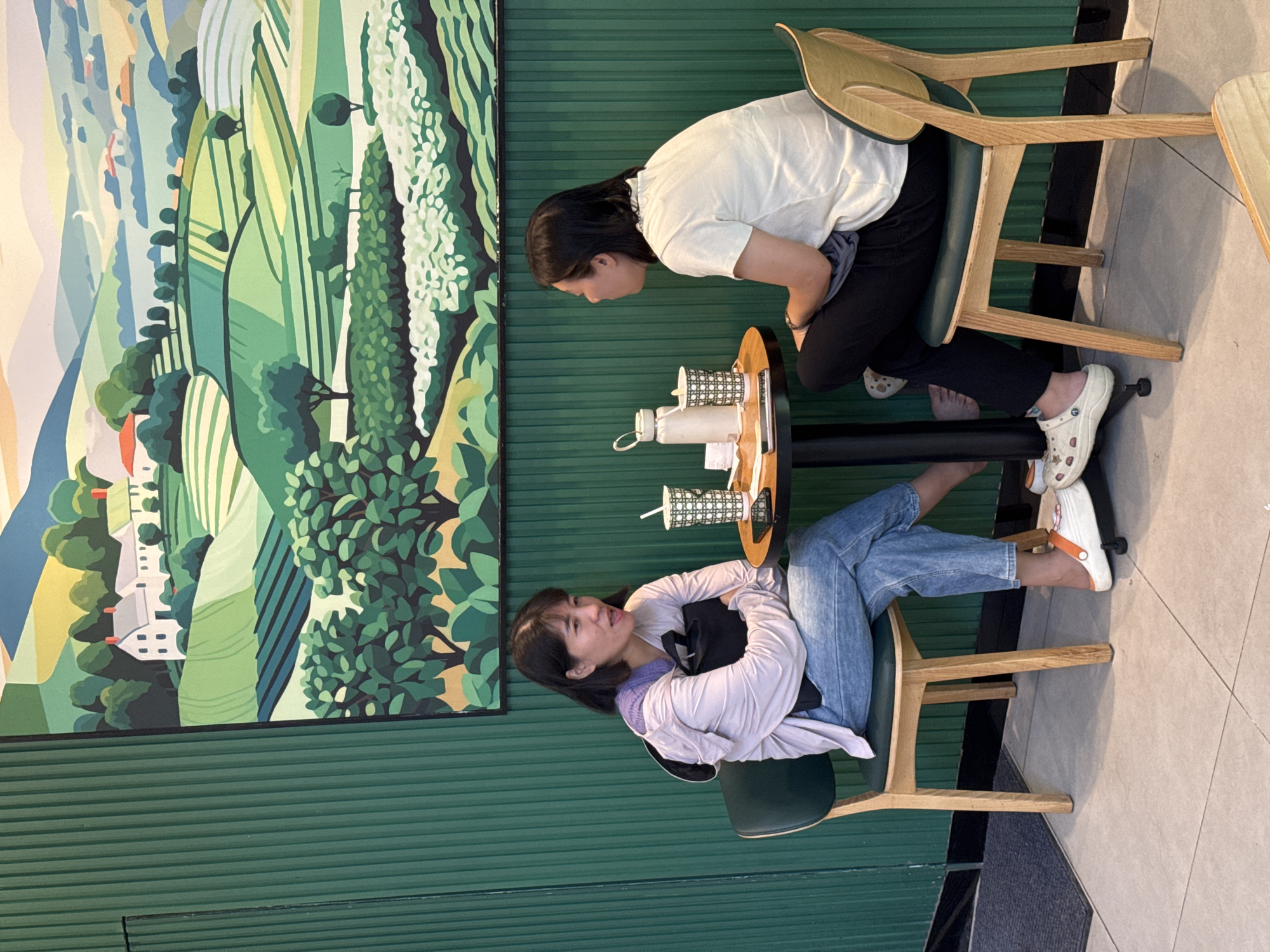 Two Asian women are seated at a small round table in a cafe. The woman on the left is wearing a light purple hoodie over a white top and blue jeans, with white and orange clogs. She is holding a black bag. The woman on the right is wearing a white t-shirt and black pants, also with white clogs. Both women appear to be engaged in conversation. On the table are two drinks, one in a patterned cup with a straw, and a white water bottle, along with a mobile phone and some papers. The cafe has green, ribbed walls and a large painting of a stylized landscape with rolling hills and houses. The flooring is tiled. The setting is indoors, likely a cafe in Nha Trang, Vietnam.