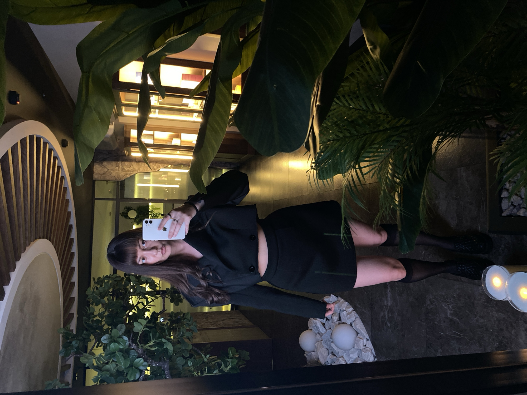 A woman is taking a selfie in a dimly lit interior space, possibly a restaurant or hotel. She's holding a white smartphone, wearing a dark short skirt and matching jacket, with black sheer stockings. The environment includes green plants, decorative white spheres on small rocks, and a partially visible wall feature of vertical wooden slats. The precise location in Votkinsk, Russia, cannot be determined from this image alone.
