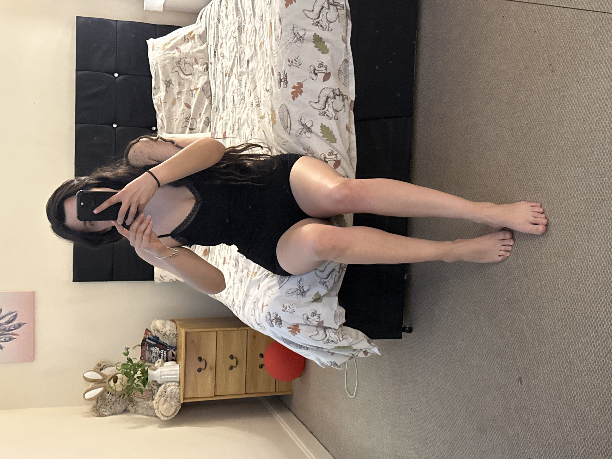 A woman is lying on a bed in a room. The bed has a black headboard and is covered with a patterned duvet. She is wearing a black top. She is holding a phone and taking a picture of herself. There is a bedside table next to the bed with a stuffed bunny and other items on it. A red balloon is next to the cabinet. The location is indicated by the style of the room and the furniture in the city of Great Yarmouth, United Kingdom.