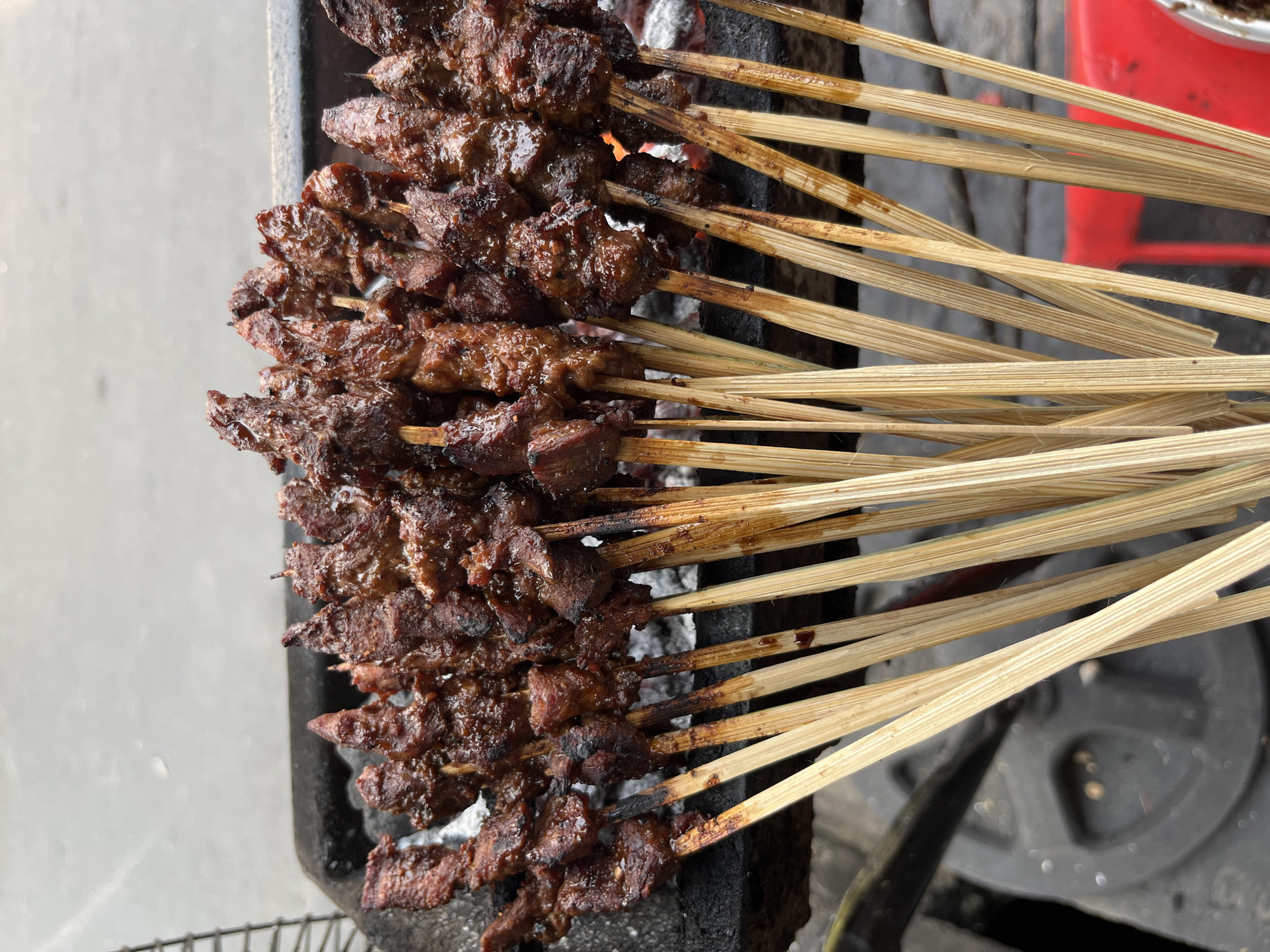 A close-up, high-angle shot shows a grill loaded with many skewers of grilled meat. The meat is dark brown, glistening with sauce, and some pieces show charring from the grill. The skewers are made of light-colored wood and are clustered together. The grill appears to be a black metal grate placed over a bed of glowing embers. In the background, a blurred grayish-white surface suggests an outdoor setting, possibly a street or pavement. To the left of the grill, a dark, textured surface, possibly a black plastic or metal object, is visible. The bottom of the image shows more wooden skewers, with some appearing slightly wet or oily. The overall scene suggests a street food vendor preparing or serving grilled meat skewers in Central Jakarta, Indonesia.