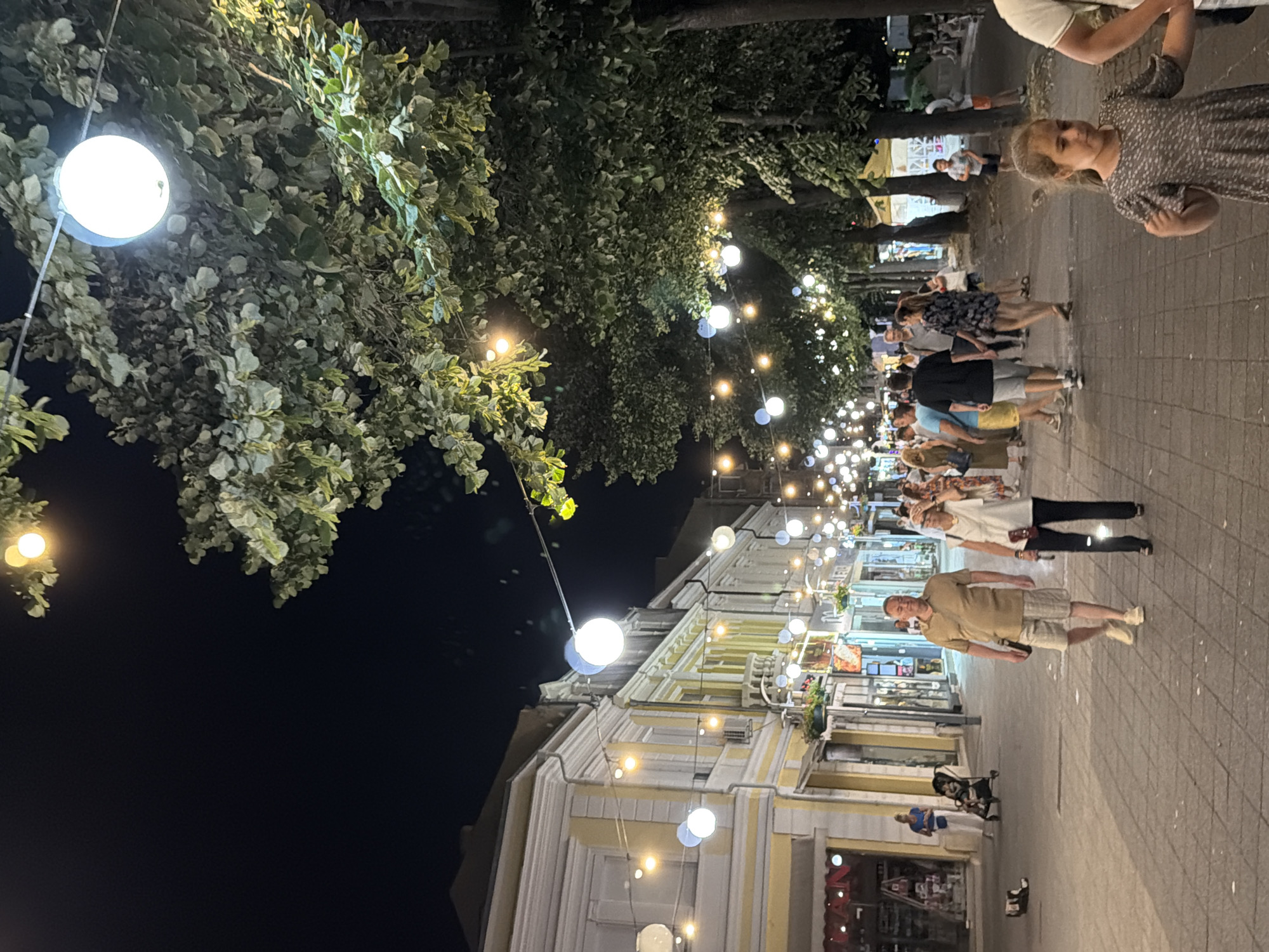A nighttime scene depicts a pedestrian street in Burgas, Bulgaria, illuminated by numerous strings of spherical lights and larger, circular hanging lamps. Trees with dense foliage line the right side of the street, their leaves catching the light. On the left, traditional European-style buildings with yellow facades and white trim house shops, their windows displaying various wares.
Several people are walking along the paved street. In the foreground, a young girl in a grey patterned dress is walking towards the right, her head tilted downwards. Behind her, a man and woman are walking away from the camera, the woman in a white top and dark pants, the man in a beige shirt and patterned shorts. Further up the street, a diverse group of individuals are strolling, some in pairs, others in larger clusters. A baby in a stroller is visible near the shops on the left, with a woman in a blue top standing beside it. Two pairs of shoes are inexplicably placed on the ground in the mid-left section of the pavement. The overall atmosphere suggests a lively evening promenade.