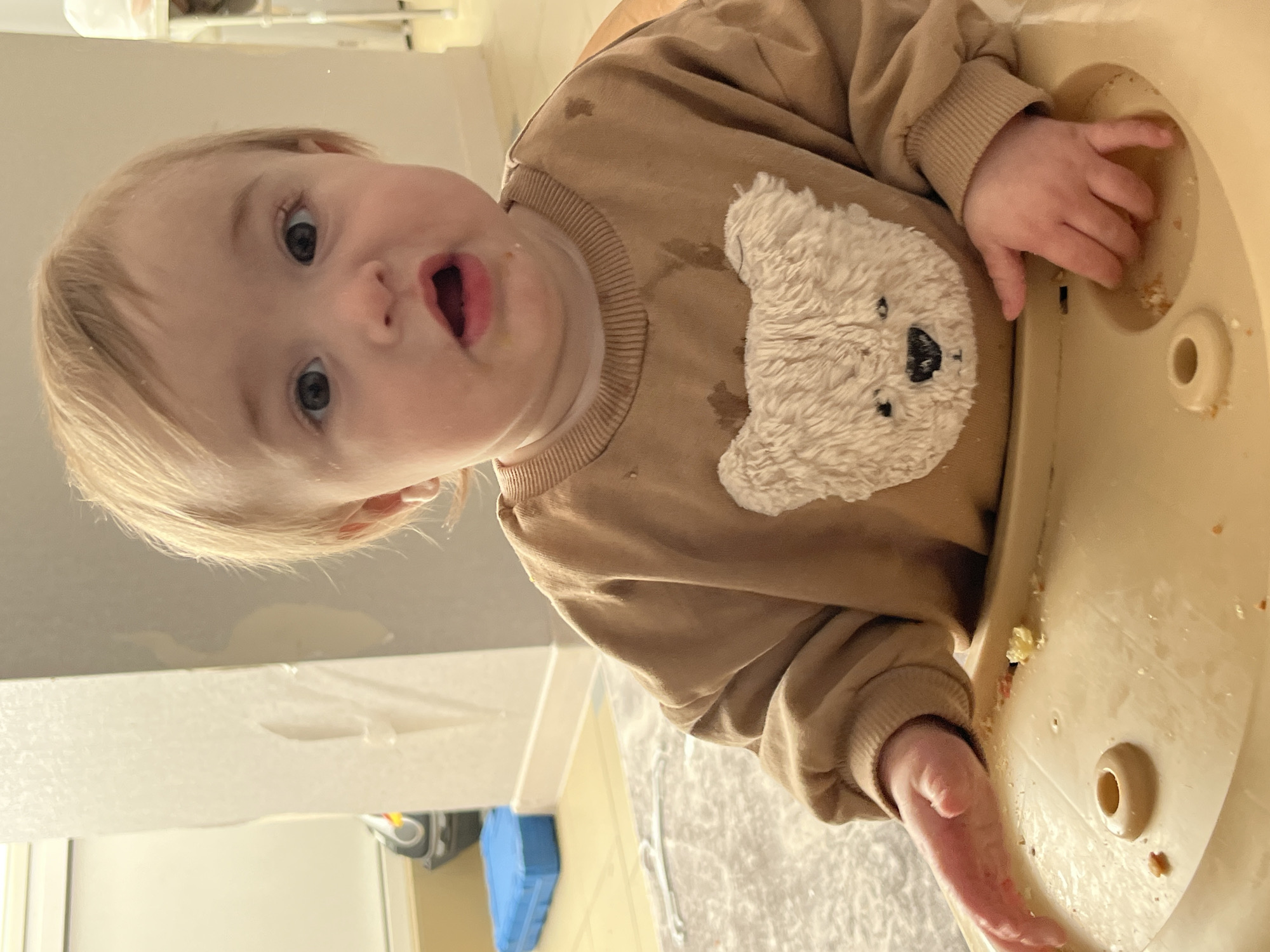 A close-up shot depicts an infant seated in a high chair. The baby has fair skin, light-colored hair, and gray eyes. The infant is wearing a brown sweatshirt with a teddy bear design on the front. The baby's mouth is open, suggesting surprise or a sound. The high chair's tray shows evidence of food. The background includes parts of a room interior, revealing a white surface and some flooring. There is no verifiable evidence in the image to place the location in Chaikovsky, Russia.
