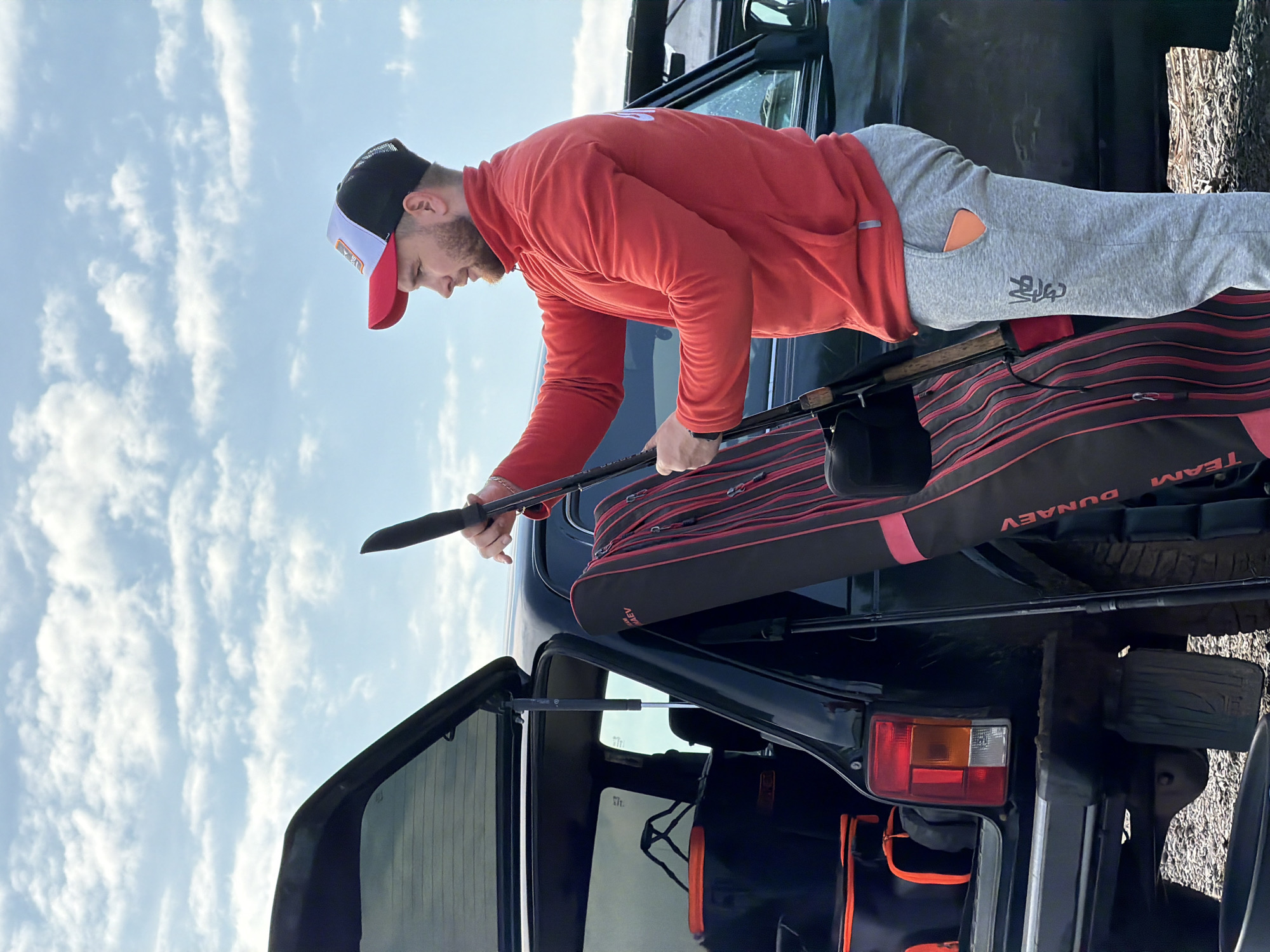 A man in a red long-sleeved shirt and grey sweatpants stands near the open trunk of a dark-colored vehicle, possibly a car or SUV. He is wearing a cap with a red bill, white front, and black mesh back. He is holding a black fishing rod, possibly preparing to store or retrieve it from a black and red bag labeled "DUNAEV," which appears to be resting on the trunk's ledge. The vehicle's trunk is open, revealing additional dark-colored bags with orange detailing inside. The backdrop shows a partly cloudy sky. The location is possibly in or near the city of Chaikovsky, Russia. The man appears to be handling fishing equipment near his vehicle.