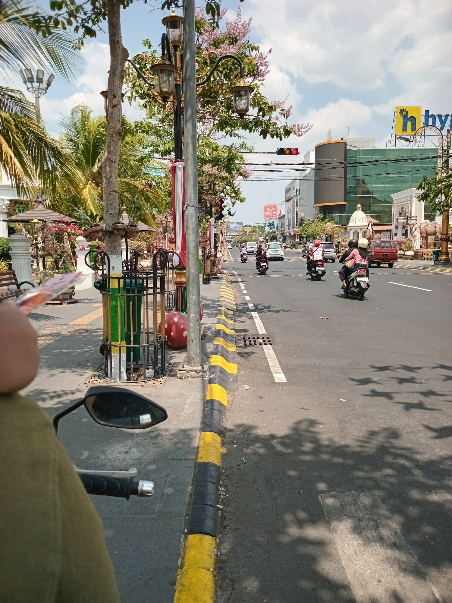 The image shows a street scene in a city with several vehicles and pedestrians. The perspective is from someone riding on a motorcycle, with the handlebars and a rearview mirror visible in the foreground. The street is paved and has yellow and black painted markings along the edge of the sidewalk.
In the background, there are several buildings, some of which appear to be commercial establishments with large windows and signage. One building has a large yellow sign with the letter "h" and stars. Trees and palm trees line the street, and there are streetlights and traffic signals visible.
Several motorcycles and cars are driving on the road. Some people are riding on the motorcycles, wearing helmets. There is a sidewalk on the left side of the road with some decorative elements, including planters and what appear to be umbrellas providing shade. The sky is partly cloudy.