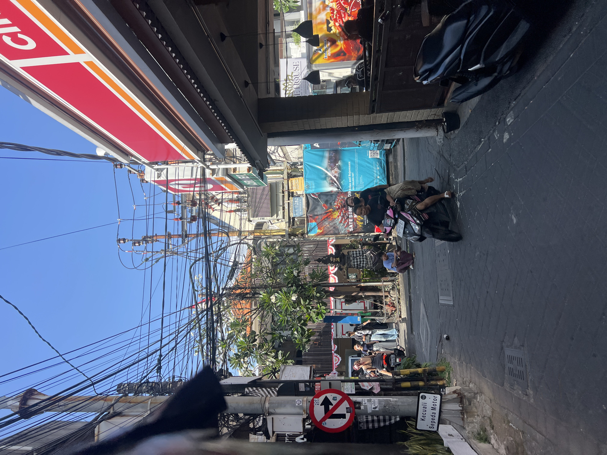 A street scene in Kuta, Indonesia, shows a narrow road lined with buildings and dense overhead electrical wires. In the foreground, a black scooter is parked. Further down the street, two people are riding a pink scooter. To the left, a sign reads "kecuali Sepeda Motor," meaning "except motorcycles," and a circular red sign prohibits right turns. Several people are walking and standing on the sidewalk. A building with a red and white facade and a large red sign is on the right, possibly a convenience store or restaurant. The sky is clear and blue, indicating a sunny day.